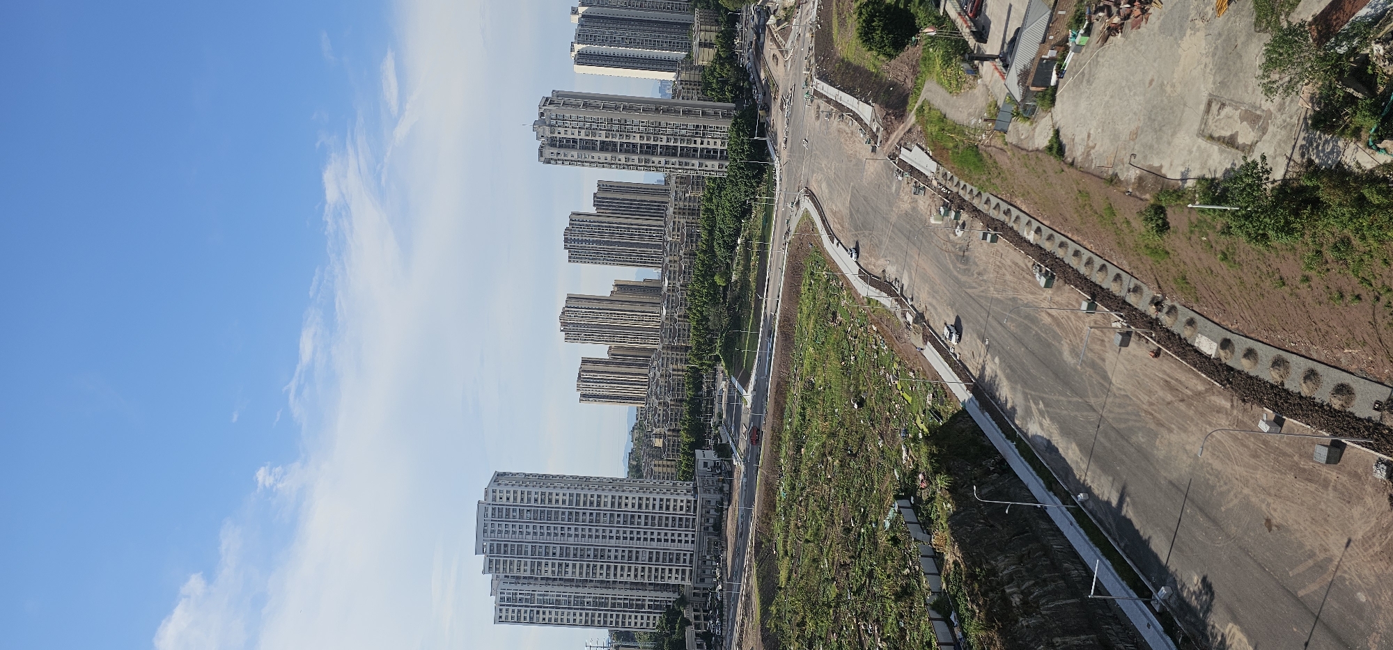 The image captures a sprawling construction site in the Jiulongpo District of China, characterized by a developing urban landscape. In the background, numerous high-rise residential buildings, appearing to be apartment complexes, dominate the skyline under a clear blue sky with scattered white clouds. The buildings vary in architectural style and height, with some featuring light-colored facades and repetitive window patterns.
In the foreground, the scene transitions to a construction zone. A wide, unfinished road network, seemingly under development, cuts through the landscape. The road surface is primarily bare asphalt or concrete, with construction materials and debris intermittently visible. Along the sides of the road, there are areas of cleared land with sparse vegetation and some piles of soil or gravel, indicating ongoing earthwork and landscaping. Rows of newly installed streetlights are present, lining the roadways.
Several white vehicles, likely construction vehicles or cars, are parked or moving along the road. A few individuals, appearing as small figures from this aerial perspective, are present on foot near the road and construction areas, engaged in various activities. The overall impression is one of significant urban development and infrastructure construction. The surrounding terrain shows some natural undulations, with patches of greenery and trees interspersed with the construction zones. The image is taken from an elevated viewpoint, providing a comprehensive overview of the area.