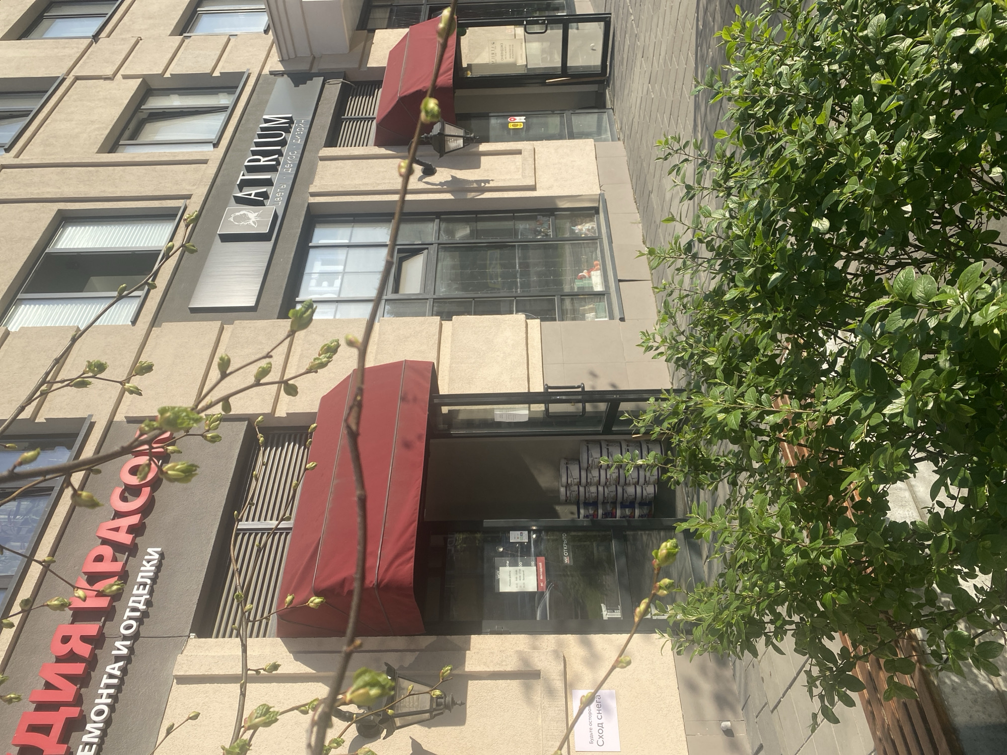 The image shows the façade of a building in Yekaterinburg, Russia. Two storefronts are visible, one is called "Atrium", with "flowers, decor, design" written beneath. The other shop is called "ДЛЯ КРАСО" (DLYA KRASO) with "ремонта и отделки" (REMONTA I OTDELKI - repair and decoration) beneath. Both storefronts have red awnings. A sign on the ground near the DLYA KRASO shop reads "Будьте осторожно Сход снега" ("Be careful, snow is falling"). There is a tree or bush on the right side of the frame, partially obscuring the view. The building is constructed of beige stone or concrete, and a sidewalk is visible in the upper right.