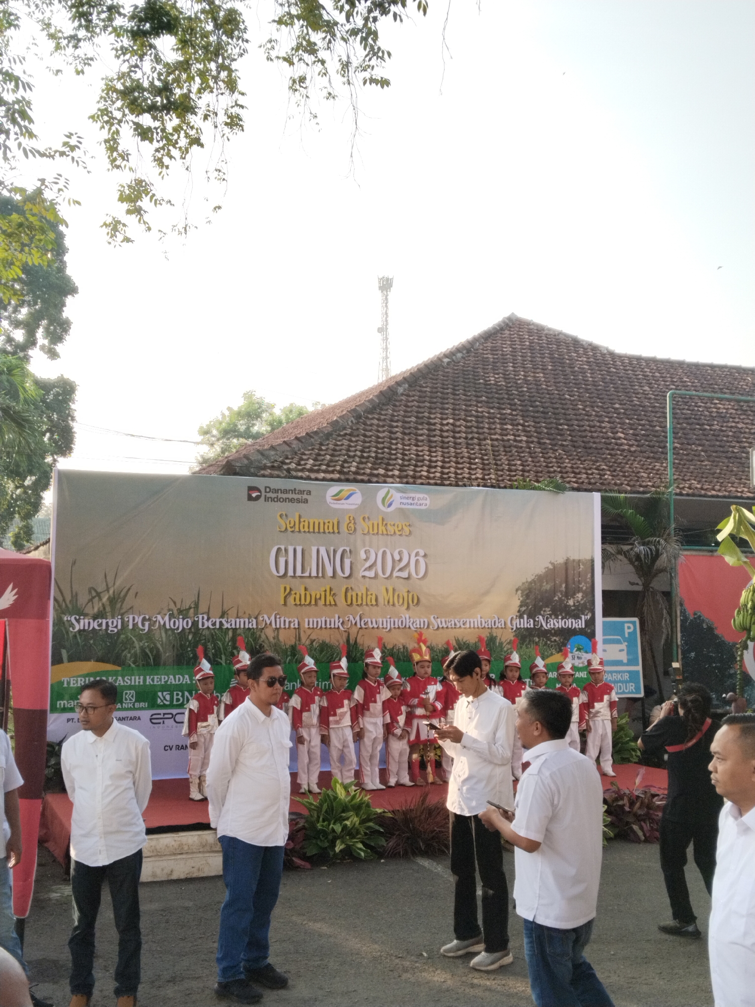 An outdoor event is depicted, likely a ceremony or celebration, possibly for the "Giling 2026" (milling season) of the Mojo Sugar Factory in Sragen, Indonesia. The scene is brightly lit, indicating daytime with a clear sky.
In the midground, a large banner is prominently displayed. It features the text "Selamat & Sukses GILING 2026 Pabrik Gula Mojo" and the slogan "Sinergi PG Mojo Bersama Mitra untuk Mewujudkan Swasembada Gula Nasional" (Synergy of Mojo Sugar Factory with Partners to Achieve National Sugar Self-Sufficiency). The banner also displays logos for "Danantara Indonesia," "sinergi gula nusantara," and "TERIMA KASIH KEPADA" (Thank You To) sponsors including "BNI," "BANK BRI," "PT. LANTARA," "EPC," and "CV RAN."
On a raised stage directly in front of the banner, a group of approximately ten children, dressed in red and white uniforms with plumed hats, stand in formation. They appear to be a marching band or drum corps, possibly performing or awaiting their turn. Potted plants are arranged as stage decorations.
In the foreground, several adult men are visible, predominantly wearing white collared shirts and dark trousers or jeans. They appear to be attendees or organizers, observing the event. One man wears sunglasses, and another is looking at his mobile phone. On the far right, a person in dark attire, possibly a photographer, is partially visible.
The background includes a building with a textured, reddish-brown tiled roof, numerous green trees, and a tall communication tower extending into the bright sky. A blue parking sign with a white "P" and the text "PARKIR MUNDUR" (Reverse Parking) is also seen on the right. The overall impression is of a structured, celebratory community or corporate gathering.