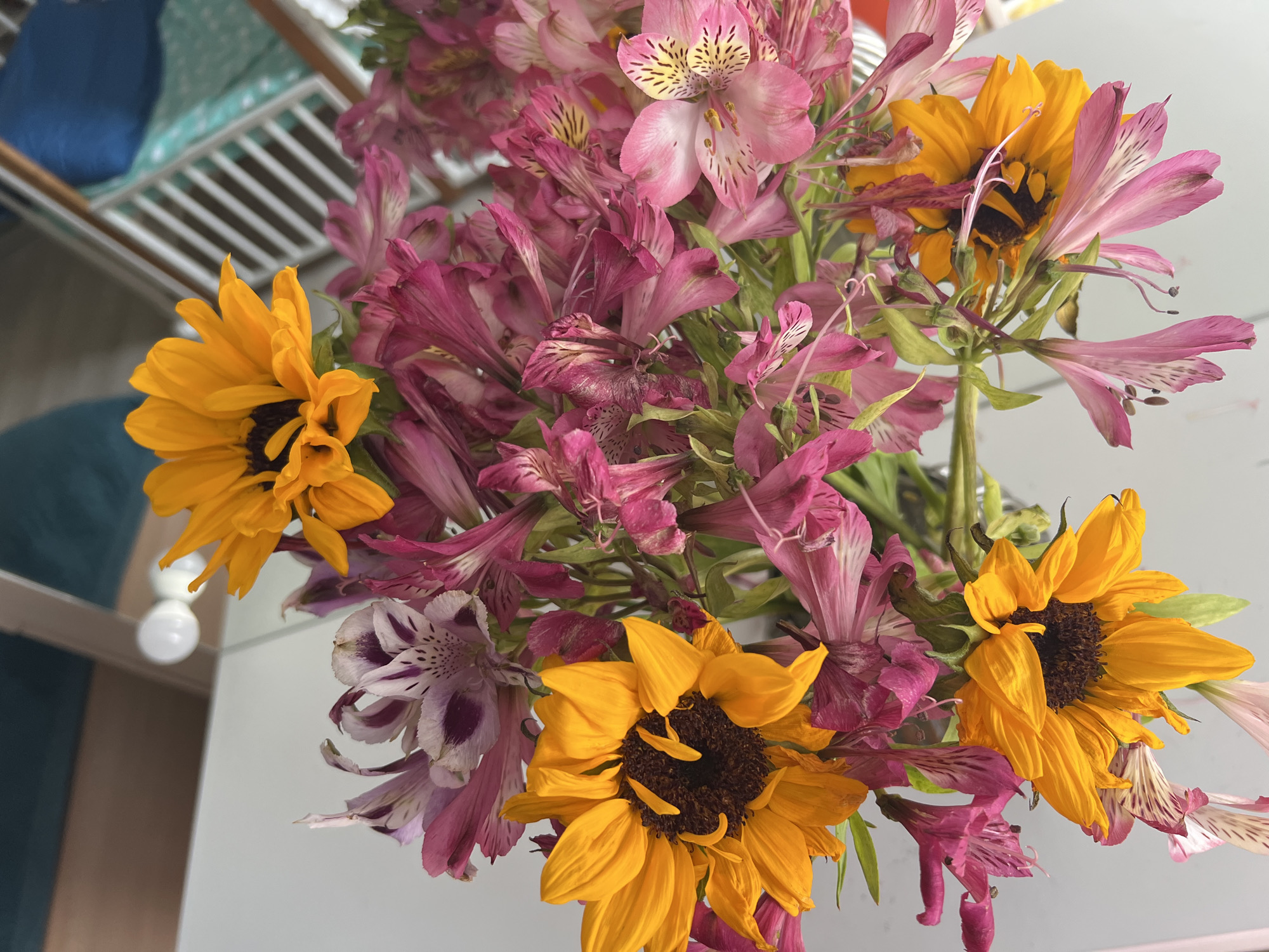 A bouquet of flowers, primarily pink alstroemeria and yellow sunflowers, is displayed in a clear vase against a blurred background. The arrangement features a mix of open blooms and some that appear slightly wilted. The lighting suggests an indoor setting, possibly near a window, with a hint of a ceiling and some light fixtures visible. The overall impression is of a natural, domestic scene. No specific location in Sochi, Russia can be determined from the image.