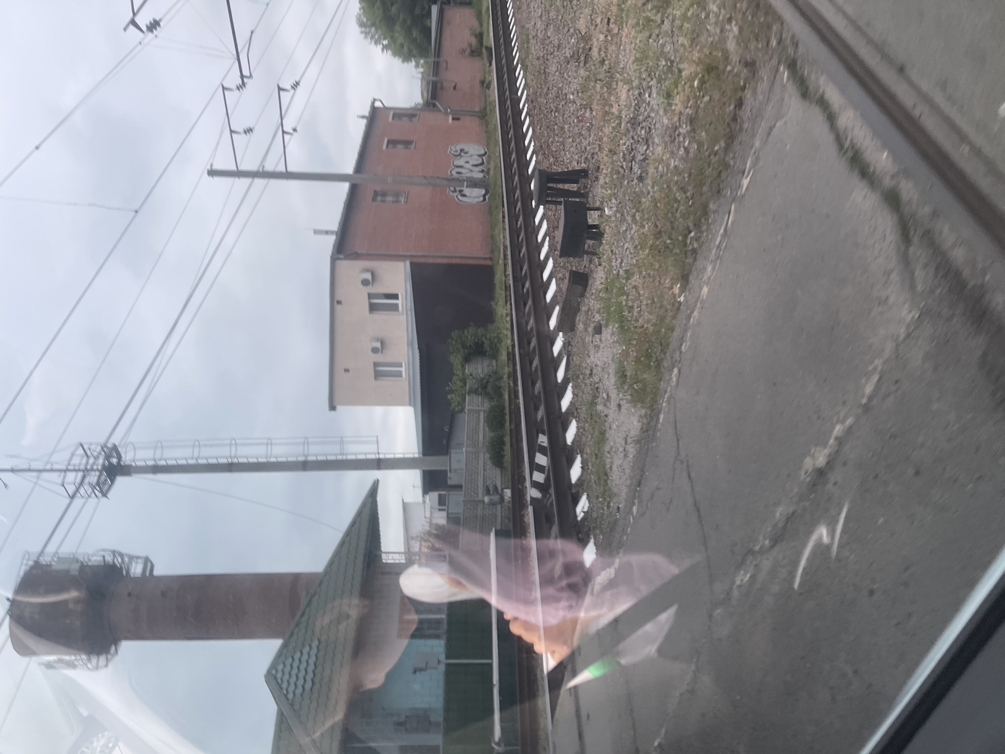 The image shows a train track with gravel and grass on the side. There are several buildings in the background, including a brick building with graffiti on it and a lighter colored building with windows and air conditioning units. A tall, slender brick water tower is visible on the left side of the image, along with a metal tower with a ladder. Overhead, power lines crisscross the sky. In the foreground, a reflection is visible on a window, showing what appears to be a person's head and shoulders with light-colored clothing, along with a portion of a car or vehicle. The scene appears to be in an industrial or semi-rural area, likely near a train station or railway line. The location is Гожули, Ukraine.