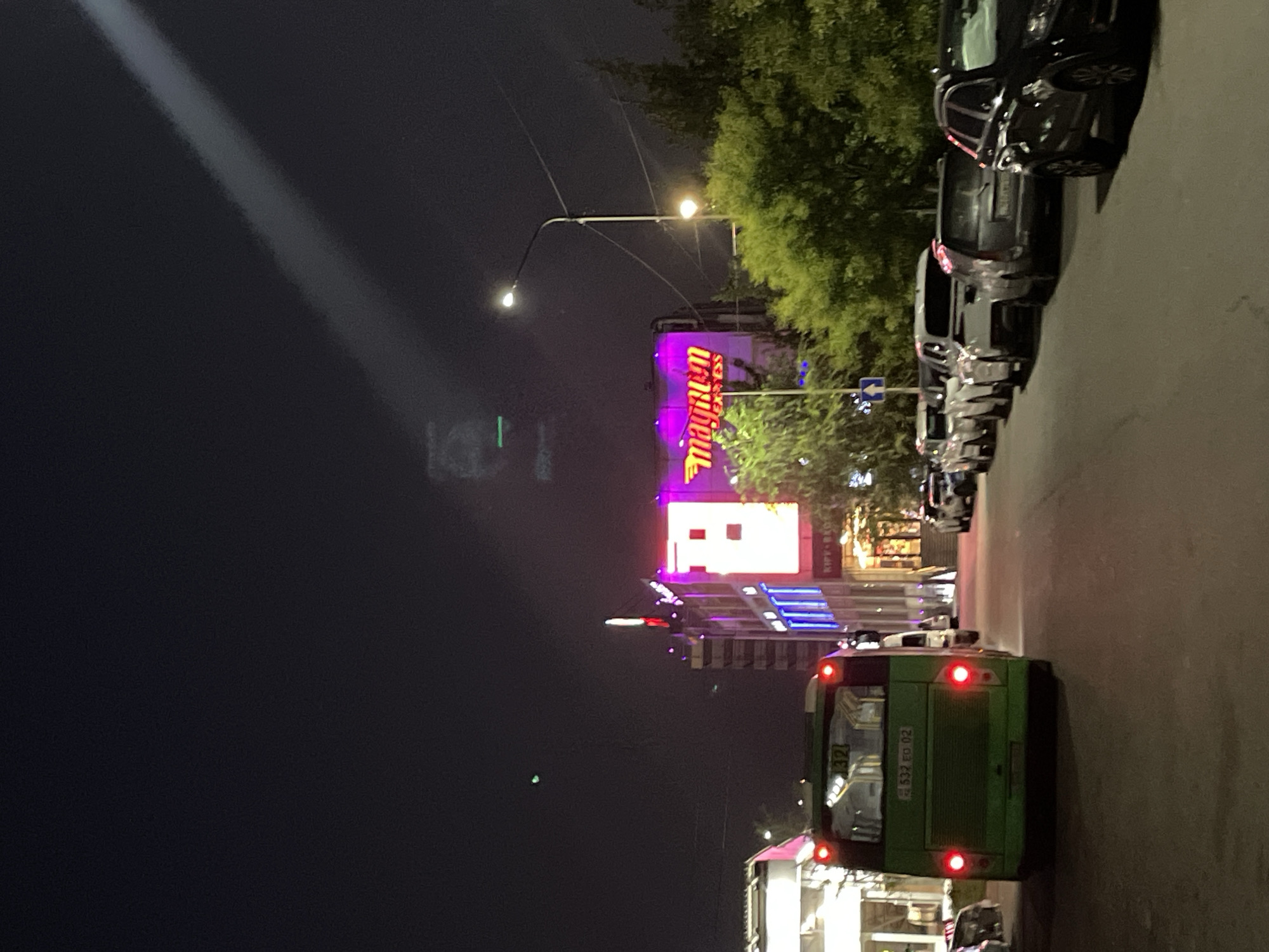 In Almaty, Kazakhstan, at night, a green bus with illuminated taillights is parked on the right side of a paved street. The bus displays the number "32" on its rear window and license plate "532 ED 02". To the right of the bus, a line of parked cars, including a black SUV in the foreground, is visible. Across the street, a building with bright pink and purple neon signage that reads "magnum EXPRESS" is illuminated. A tall street lamp with a glowing bulb stands near the building. Trees with green foliage are situated behind the building, and a faint green light is visible in the dark sky. A bright white beam of light cuts across the night sky from the upper right portion of the image.