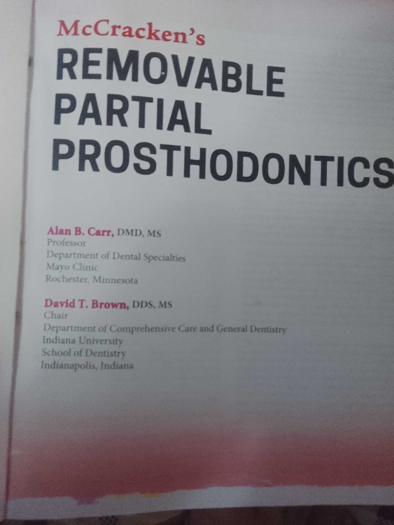 This media file is a photograph of an open book, likely a textbook or publication, displaying the title page or a similar introductory section. The prominent text at the top reads "McCracken's REMOVABLE PARTIAL PROSTHODONTICS," indicating the subject matter. Below the title, affiliations and names of individuals are listed.
The first individual featured is Alan B. Carr, identified as a DMD, MS, and Professor in the Department of Dental Specialties at Mayo Clinic in Rochester, Minnesota. The second individual is David T. Brown, a DDS, MS, who holds the position of Chair in the Department of Comprehensive Care and General Dentistry at Indiana University's School of Dentistry in Indianapolis, Indiana.
The page itself appears to be a clean, white, printed surface. The lighting suggests an indoor environment, likely a room where the book is being displayed or read. There are no discernible details regarding the time of day or weather. The overall impression is professional and academic, given the content and format. The image captures a close-up view of the text, with a slight gradient of red and pink hues appearing at the bottom of the frame, possibly from the surrounding environment or the book's cover art, though the primary focus remains on the text.