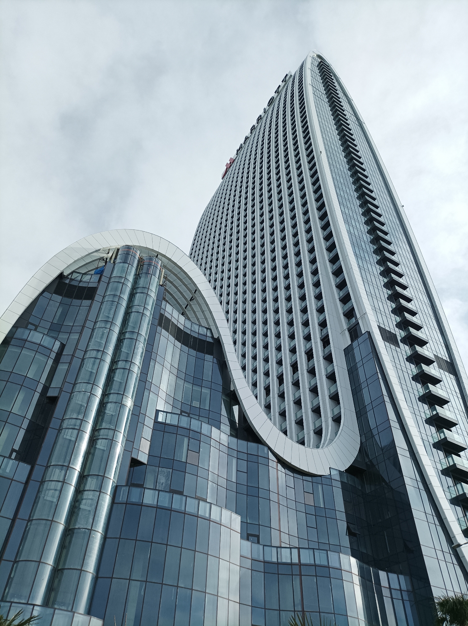 The image depicts a low-angle view of the Batumi Tower, located in Batumi, Georgia, specifically identified by the visible "MARRIOTT" signage on its upper facade. The scene is dominated by two interconnected modern architectural structures under an overcast sky.
The taller structure is a sleek, multi-story skyscraper, characterized by a predominantly glass and light-grey facade that curves and tapers upward. Its left side displays numerous regularly spaced, dark-framed windows, while the right side features vertical rows of recessed balconies or external design elements. The word "MARRIOTT" is horizontally aligned near the building's apex on the left, accompanied by a small red logo element.
Connected to the base of the skyscraper is a distinct, lower multi-story building. This structure features a prominent, undulating white or light-grey canopy or roofline that creates a wave-like appearance. Its facade is composed largely of dark-tinted glass panels. Two cylindrical, glass-enclosed structures, possibly external elevators or decorative tubes, are visible beneath the wavy canopy.
The overall environment is primarily architectural, with only faint green foliage visible at the very bottom of the frame, suggesting an outdoor urban setting. No people, discernible actions, or interactions are visible within the image.