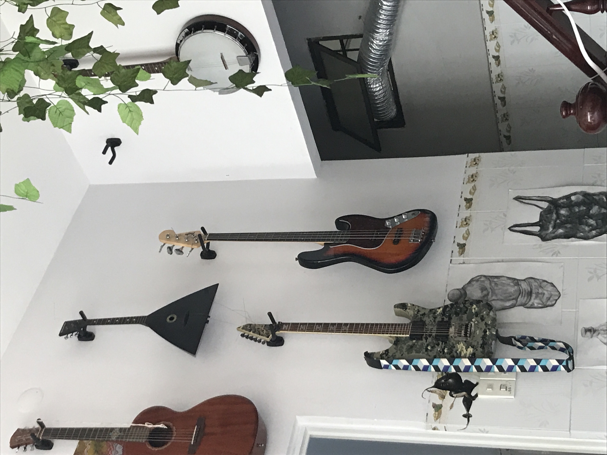 The image shows a collection of musical instruments mounted on a white wall. From top to bottom, there is a banjo, a bass guitar, a balalaika, an electric guitar with a blue and white strap, and an acoustic guitar. Green decorative vines are draped near the banjo. The wall is decorated with painted tiles that appear to be dog themed. A vent is visible in the upper center of the image. The location is consistent with the city of Nha Trang, Vietnam. There are no people visible in the image.
