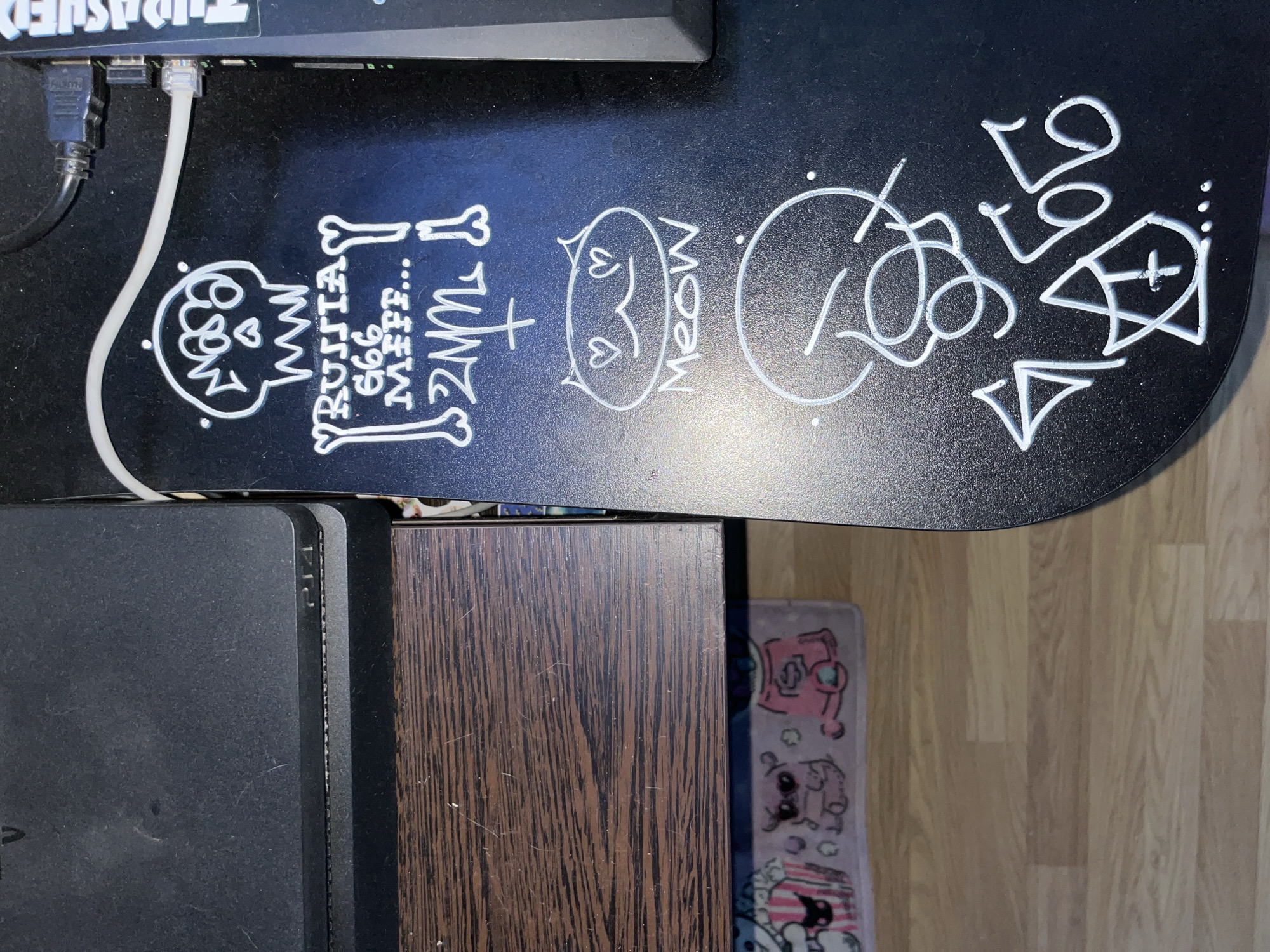 The image shows a close-up view of a black surface, likely a desk or piece of furniture, with white graffiti markings. The graffiti includes a skeleton head, text that reads "RUSSIA 666 MEFF...", a drawing of a cat face with hearts for eyes and the word "Meow" below it, and other abstract symbols and numbers, including "666" and a stylized cross. A portion of a black PlayStation console is visible in the upper left corner. Electrical cables are plugged into a device on the right side of the frame. The surface appears to be slightly dusty. The background is blurred and shows a wooden floor and what appears to be a mat with cartoonish figures. The scene is indoors, with no indication of a specific city or location.