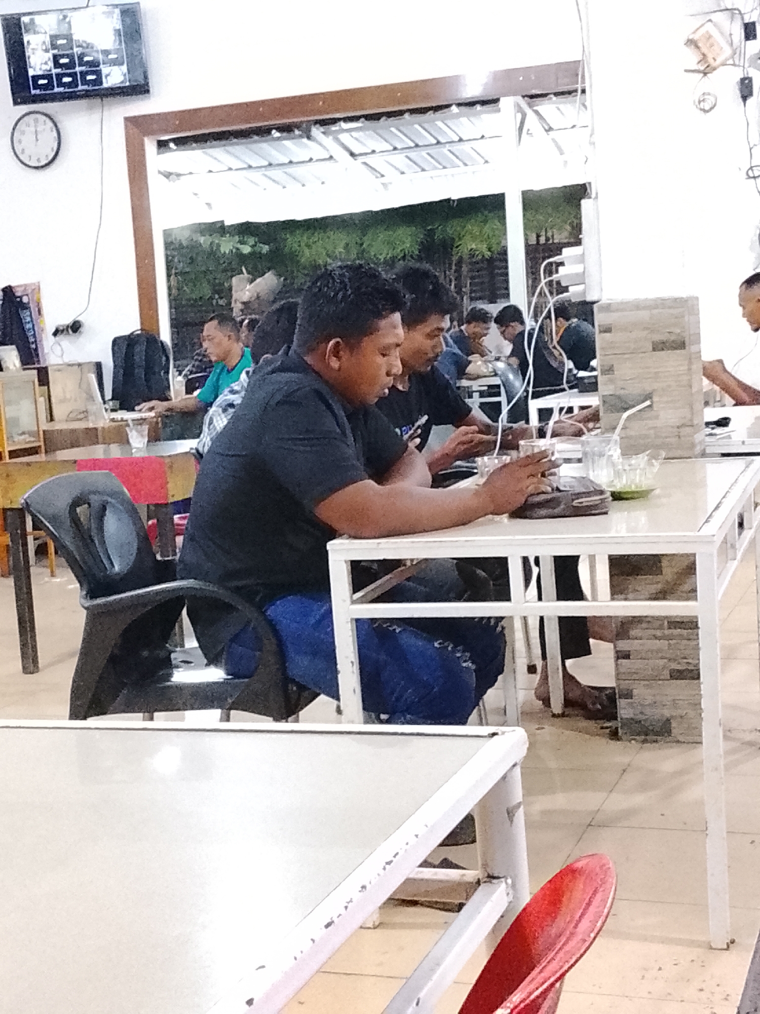 The image shows a group of men seated at tables in what appears to be an internet cafe or similar establishment. The cafe is located in the city of Lhokseumawe, Indonesia, as indicated by the location information. The men are focused on screens or devices, with some eating or drinking. Tables and chairs are arranged in rows, and the interior is brightly lit. A television is mounted on the wall, and multiple power outlets and wiring are visible, suggesting the technological nature of the space. Several tables have beverages and food, indicating a social element to the gathering. Some patrons are wearing casual clothing. One of the men is wearing blue jeans.