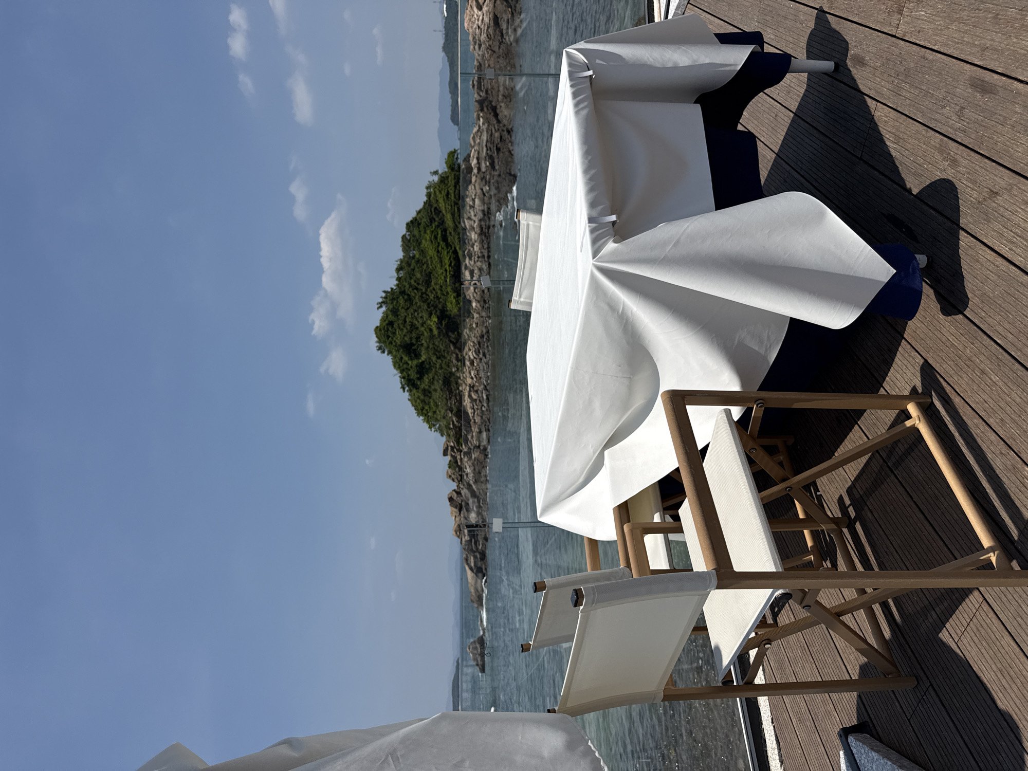 A dining table covered with a white tablecloth is set on a wooden deck overlooking a body of water. Two white canvas chairs with wooden frames are positioned in front of the table. In the background, a small, green island with rocky shores is visible, with more islands and hills in the distance. The sky is clear blue with a few scattered clouds. The scene suggests an outdoor dining area, possibly a restaurant or a private residence, in a coastal or lakeside location. The presence of a table and chairs implies a setting for dining or relaxation. The natural elements like the water and island contribute to a serene and scenic environment. It is not possible to definitively determine the exact location within Longgang District, China, based solely on the visual information provided in the image.