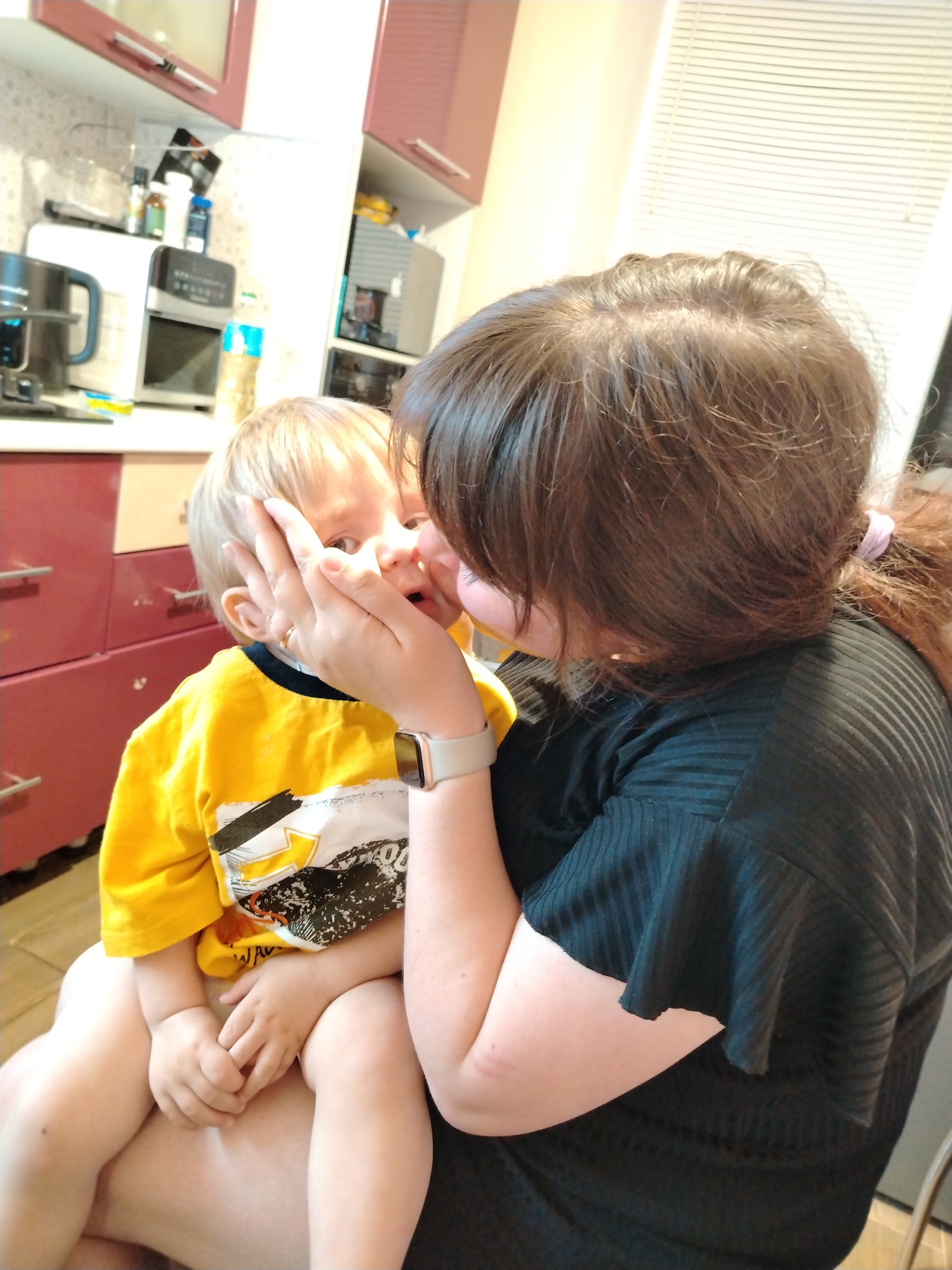 A woman is holding a young child on her lap in a kitchen environment. The child is fair-skinned, has blonde hair, and is wearing a bright yellow shirt with a print on the front. The woman has dark hair pulled back in a ponytail and is wearing a black short-sleeved shirt. She is holding the child's face with her hand. The kitchen has red and white cabinets. A coffee maker and microwave are visible on the counter. Venetian blinds are seen in the background. Based on the information provided and general knowledge, it is impossible to confirm if the image was taken in the city of Noviy, Russia.