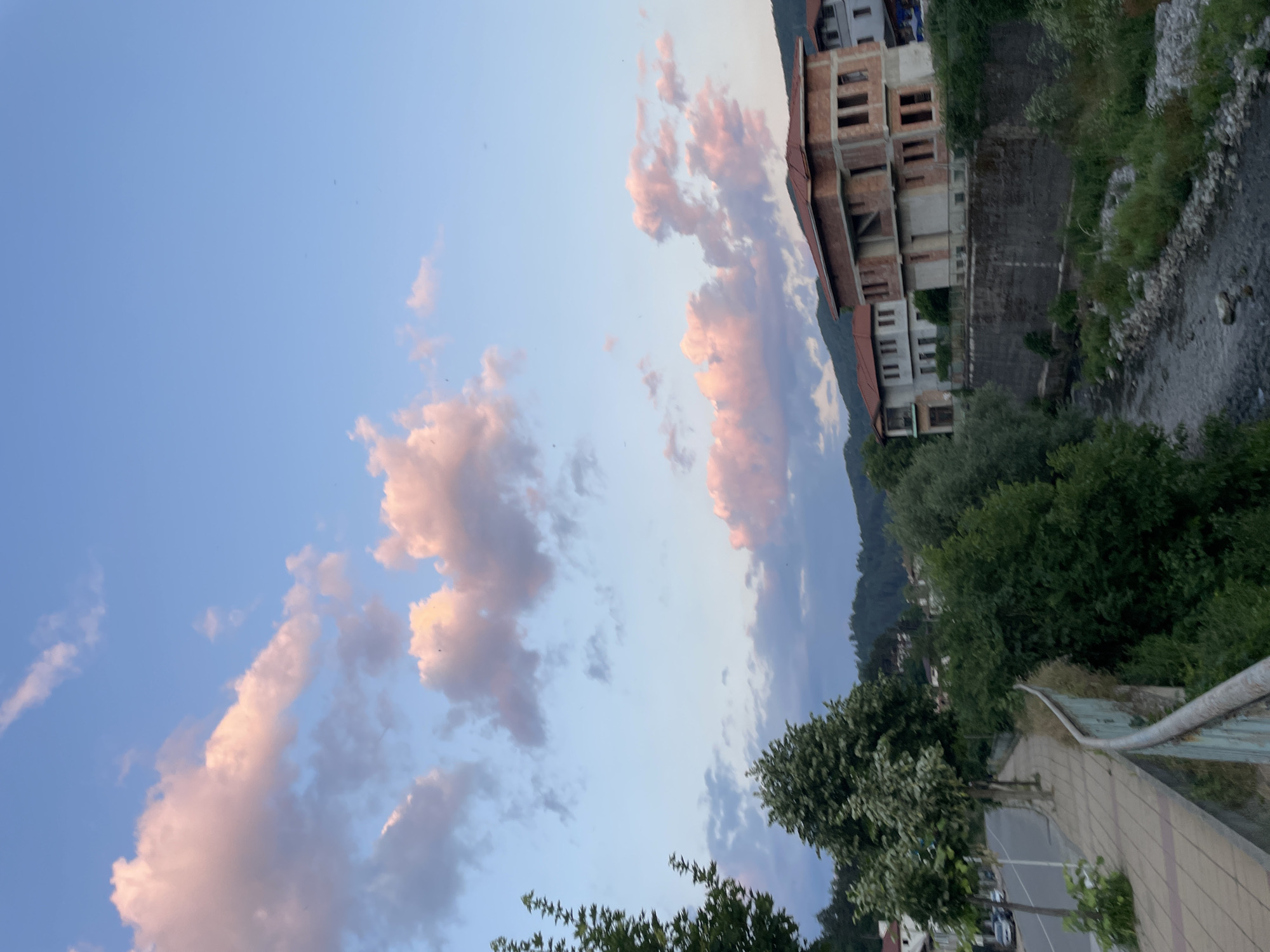 The image depicts a scenic view of Zlatograd, Bulgaria, at dusk. The sky is a gradient of soft blues and pinks, adorned with scattered clouds catching the last rays of sunlight. In the background, a partially constructed building stands tall, its brickwork exposed, suggesting ongoing construction or renovation. Adjacent to the building, a river flows, its banks lined with lush green trees and rocky terrain. The foreground shows a paved pathway with a metal railing, hinting at a viewpoint or walkway overlooking the scene. The overall atmosphere is tranquil and serene, with the soft twilight colors creating a peaceful ambiance. There is no visible text in the image.