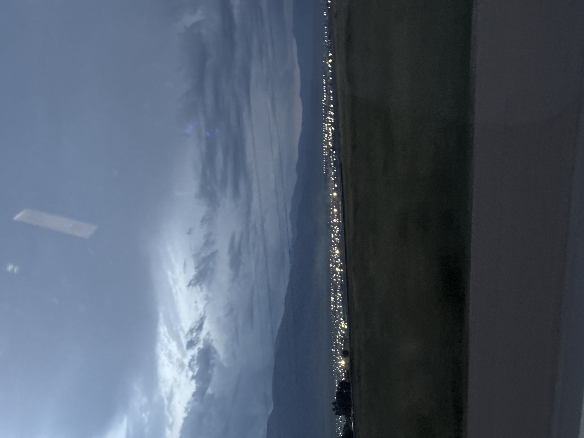 An exterior, wide shot captures a twilight view of Balykchy, Kyrgyzstan, as seen from a distance. The sky is a hazy blend of blues and grays, with indistinct clouds and a faint reflection from what appears to be a window on the left side. In the middle ground, a vast expanse of lights stretches horizontally across the scene, indicating the presence of a town or city. These lights are a mix of yellow and white, suggesting streetlights and building illumination. The town is nestled at the base of a mountain range, which forms a dark, undulating silhouette against the sky. In the foreground, a dark, grassy field occupies the bottom half of the image, with a darker strip of land or road at the very bottom. A lone tree stands on the left side of the field, near the beginning of the lit-up area. The overall impression is one of a city awakening or winding down as dusk settles.