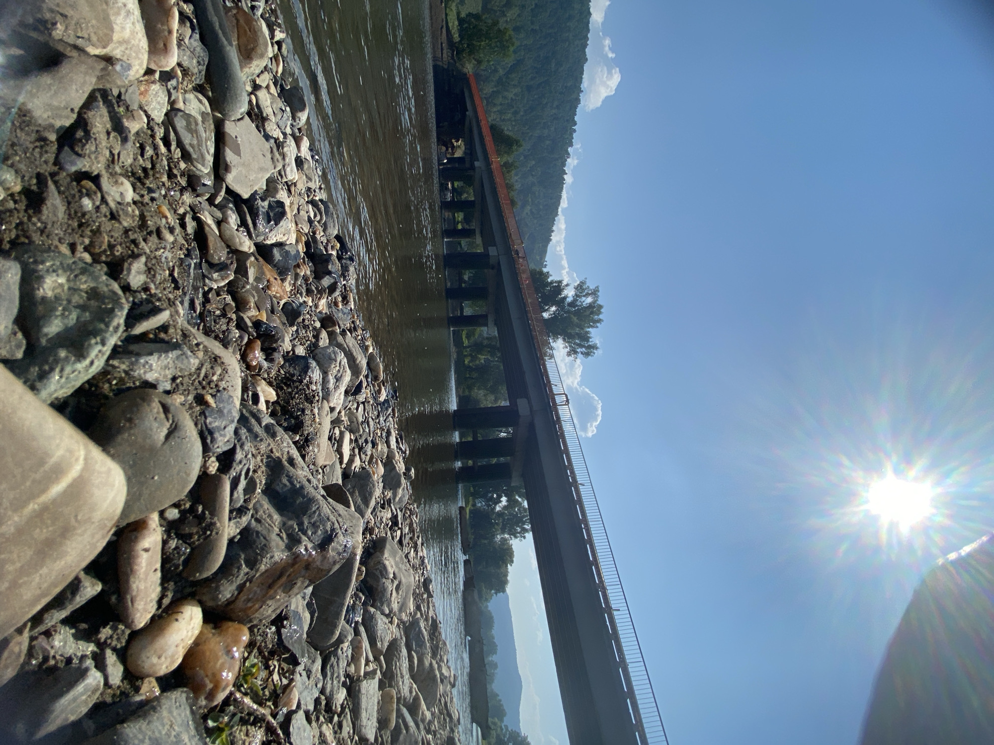 The image captures a wide-angle view of a bridge spanning a river on a clear, sunny day. The foreground is dominated by a rocky riverbank, with various sizes and colors of stones scattered across the ground. The river itself is calm, with gentle ripples reflecting the bright sunlight.
In the mid-ground, the bridge is a prominent feature, supported by multiple concrete pillars. The bridge has a railing along its length and appears to be made of concrete and metal. Trees line the banks of the river on both sides, and a forested hill rises in the background, indicating a natural or semi-rural environment. The sky is a vibrant blue, with the sun intensely bright and creating a lens flare effect in the upper right portion of the image. There is no visible presence of people or any specific urban infrastructure to suggest it is in the city of Maloarslangulovo, Russia.