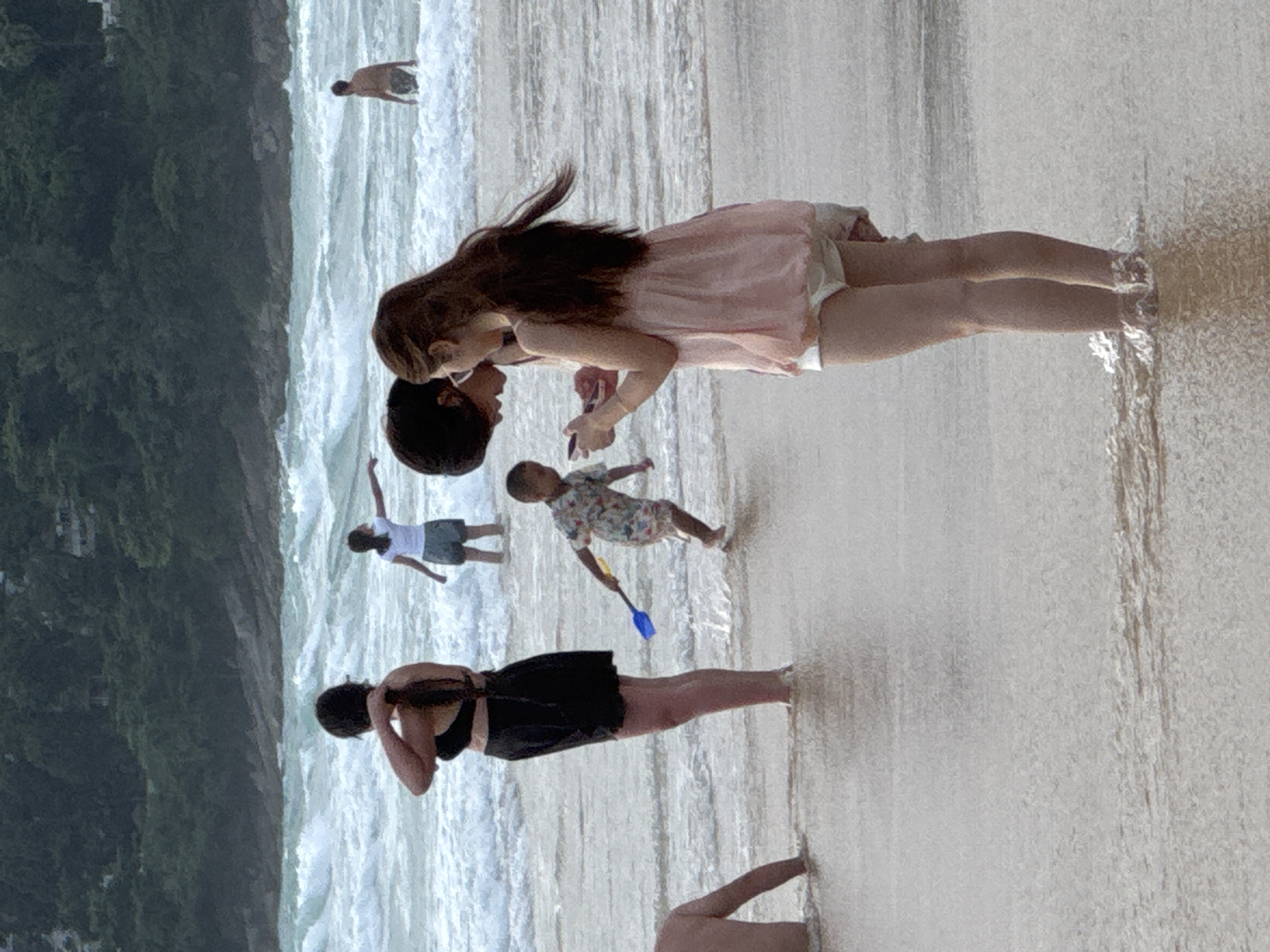 The media file depicts a beach scene at Karon, Thailand. Several people are visible, some in the water and others on the wet sand near the shoreline. In the foreground, a woman wearing a black swimsuit is standing with her arms behind her head. To her left, a person is partially visible with their legs extended.
Further down the beach, a woman in a light pink dress is leaning down towards a person who appears to be a child. The child is wearing a patterned shirt and holding a blue shovel. Behind them, another child in a white top and shorts is standing. In the background, a person is visible in the ocean. The water is foamy and appears to be waves breaking on the shore.
The overall impression is of a casual day at the beach with families and individuals enjoying the water and sand. The lighting suggests it may be daytime, possibly late afternoon given the soft light. The lush green foliage on the hillside in the background indicates a tropical location. No text is visible in the media file.