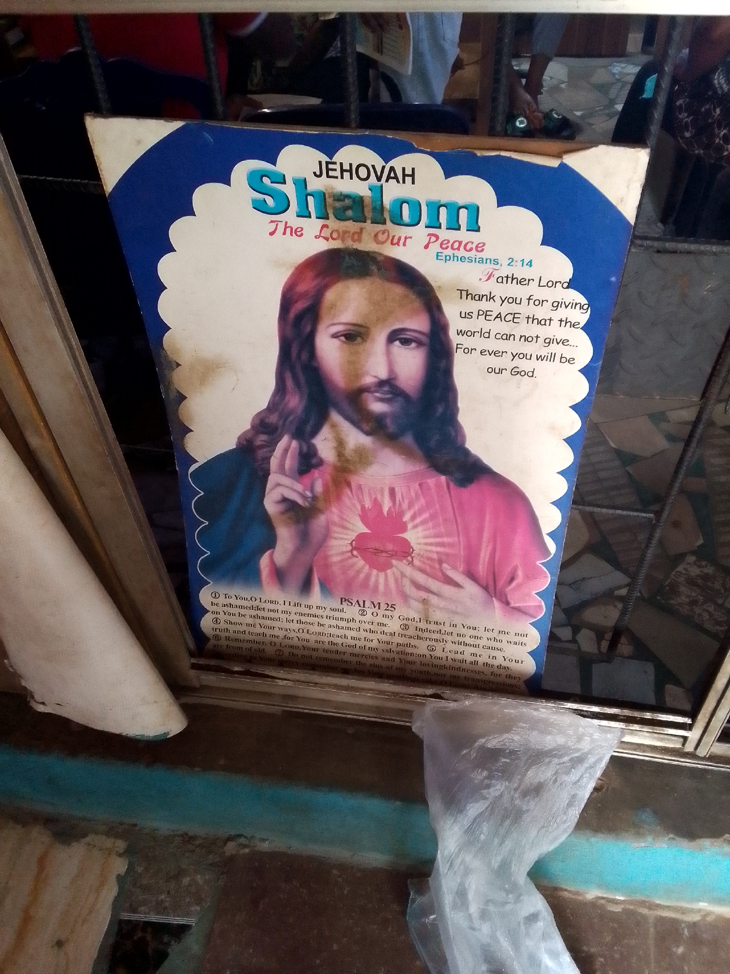 A faded poster featuring a depiction of Jesus Christ is displayed at eye level, leaning against a metal grate. The poster has a scalloped blue and white border and reads "JEHOVAH Shalom The Lord Our Peace" at the top. Below this, a prayer to "Father Lord" is inscribed, followed by the text of Psalm 25. Jesus is shown with a heart radiating light on his chest and his right hand raised in a blessing gesture. The poster is located outside on a street in Nkpor, Nigeria. The background includes a metal fence, a dark wall, and a glimpse of people in the distance. The foreground features a plastic bag lying on a blue and brown tiled surface.
