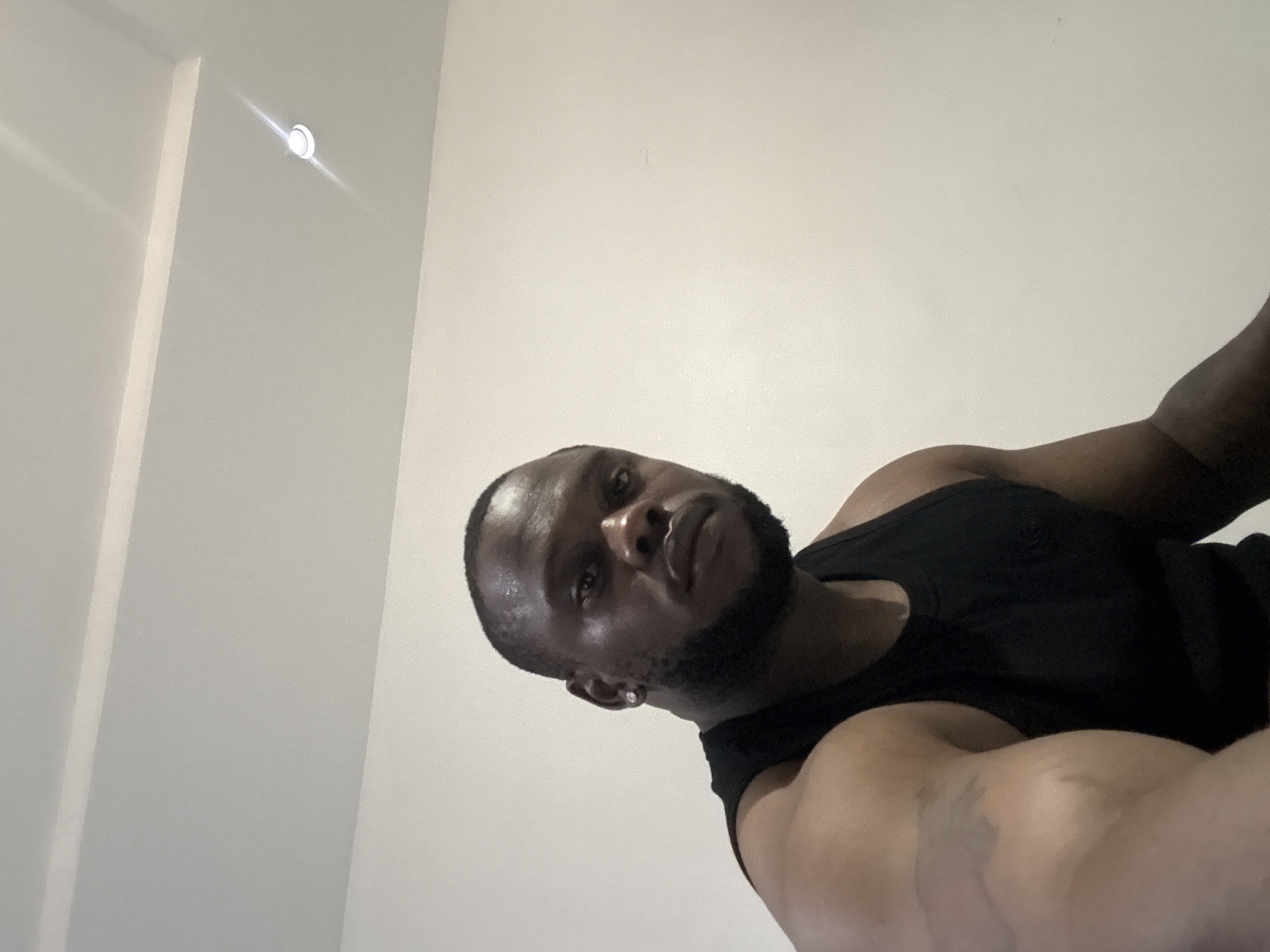 A medium shot depicts a dark-skinned man wearing a black tank top. He has a shaved head, a short beard, and an earring in his left ear. The background is a plain white wall with a recessed light fixture. The location is unknown.