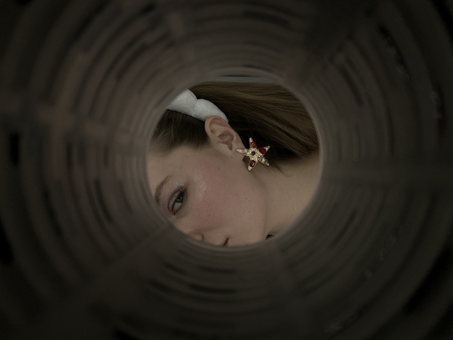 Through a cylindrical opening, a woman's face is visible. She wears a white, fluffy headband and a star-shaped earring adorned with colored gems. The background is out of focus, drawing attention to the subject's face, which is subtly illuminated. The unique perspective creates an intriguing, almost voyeuristic, feel.