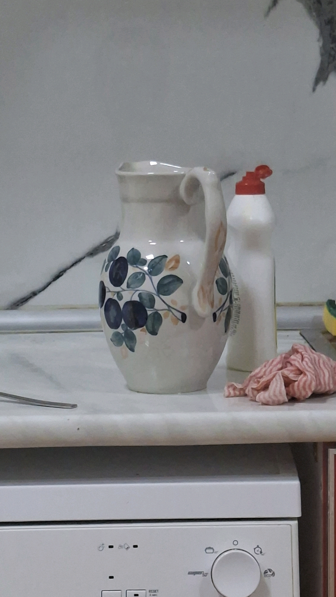 A white pitcher with a floral pattern is on a countertop in what appears to be a kitchen. To the right of the pitcher is a white bottle with a red cap, likely dish soap. To the right of the bottle, a pink cloth is crumpled. In the background, a tiled wall is visible. In the foreground, the top of a white appliance, possibly a dishwasher, with control buttons and a dial is present. This scene is located in Mingachevir, Azerbaijan.