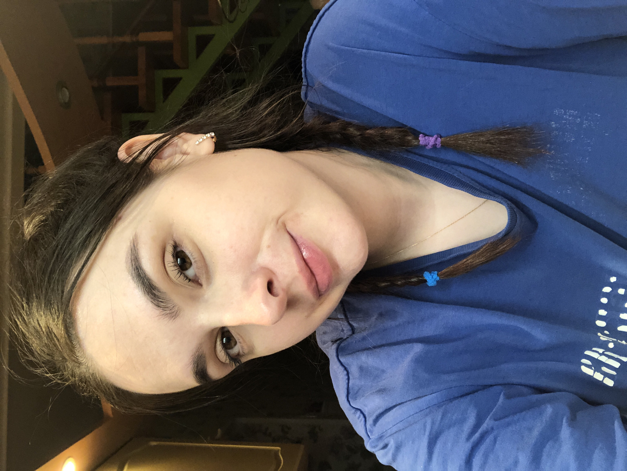 The image displays a close-up, head-and-shoulders portrait of a female subject taken indoors in the city of Noviy, Russia. The subject has fair skin, dark brown hair parted down the middle and styled into two braids; the braid on the viewer's left is secured with a blue elastic, and the braid on the viewer's right is secured with a purple elastic. She has brown eyes, full dark eyebrows, and a slight, closed-mouth smile. She wears a blue, short-sleeved t-shirt with faded white graphic print on the chest, a thin gold-colored necklace chain, and a gold-toned hoop earring featuring multiple clear stones in her right ear. The background is out of focus, showing a dark wooden structure or cabinetry with an illuminated warm light source to the upper left, and a portion of a wooden staircase or railing with a circular ceiling light fixture to the upper right.