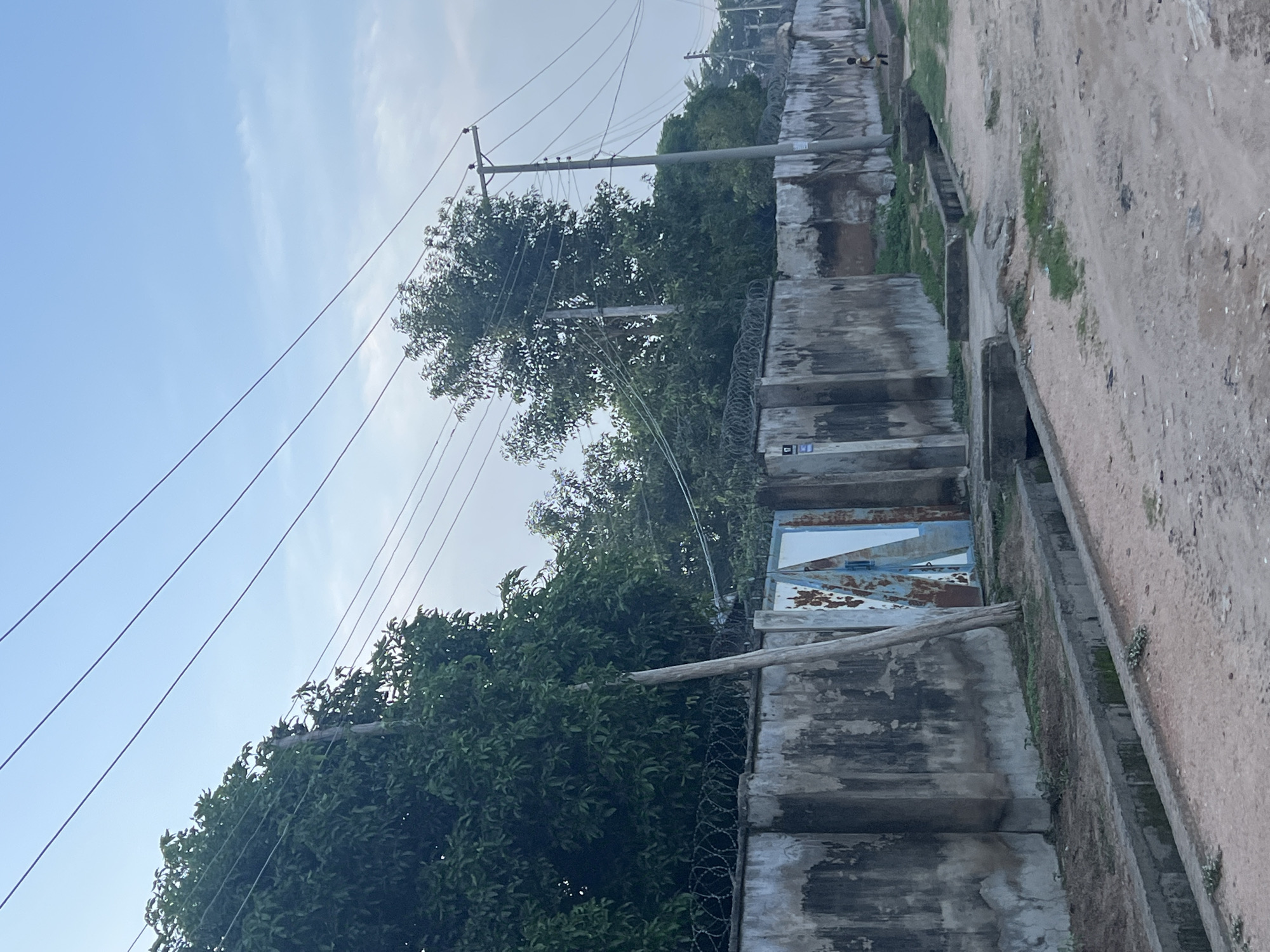 The image depicts a street scene in Jalingo, Nigeria. A weathered concrete wall, topped with barbed wire, runs along the right side of the street. Behind the wall, dense green foliage from trees obscures the view. To the left of the wall, a dirt road is visible, with a concrete drainage channel running alongside it. A rusted metal gate with white panels is partially visible in the wall. Utility poles and power lines stretch across the sky, which is a pale blue with wispy clouds. In the distance, a child is seen walking on the street. The overall impression is of an ordinary urban environment in Jalingo.