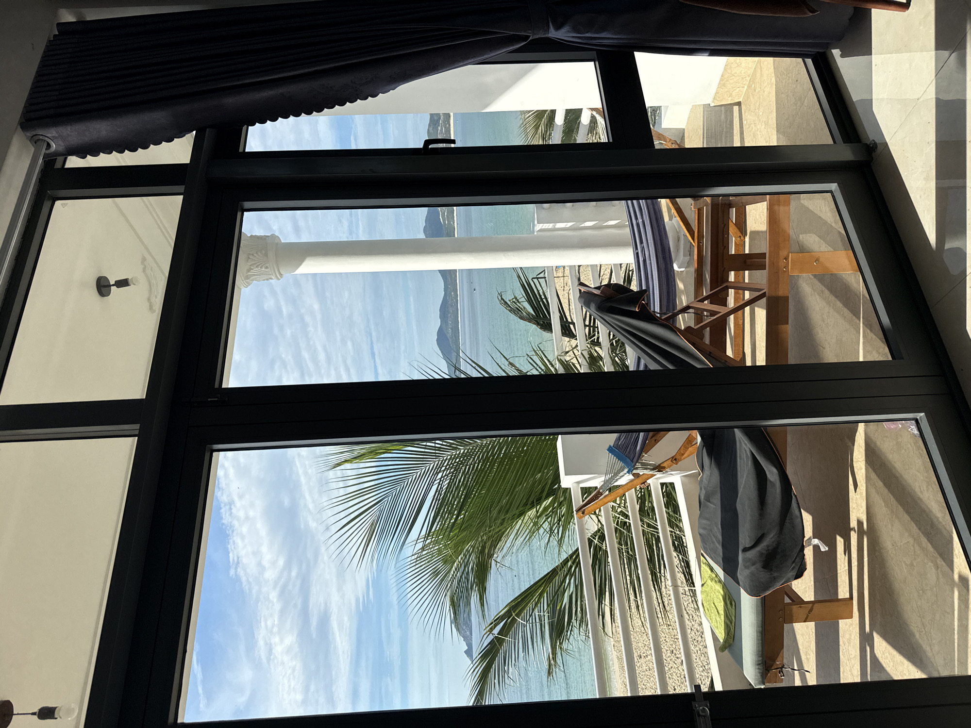 A view through a large window shows a balcony with two pieces of furniture and a beautiful ocean and beach view in Song Cau, Vietnam. On the balcony, a wooden lounge chair is covered with a black blanket and a green towel. Behind it, a hammock is suspended between two wooden frames. The balcony overlooks a white railing, a sandy beach, and the turquoise ocean. Palm fronds from a nearby tree are visible in the foreground. The sky is blue with scattered white clouds. A white pillar, part of the building's architecture, is also visible. The scene suggests a serene and relaxing atmosphere, typical of a tropical vacation destination.