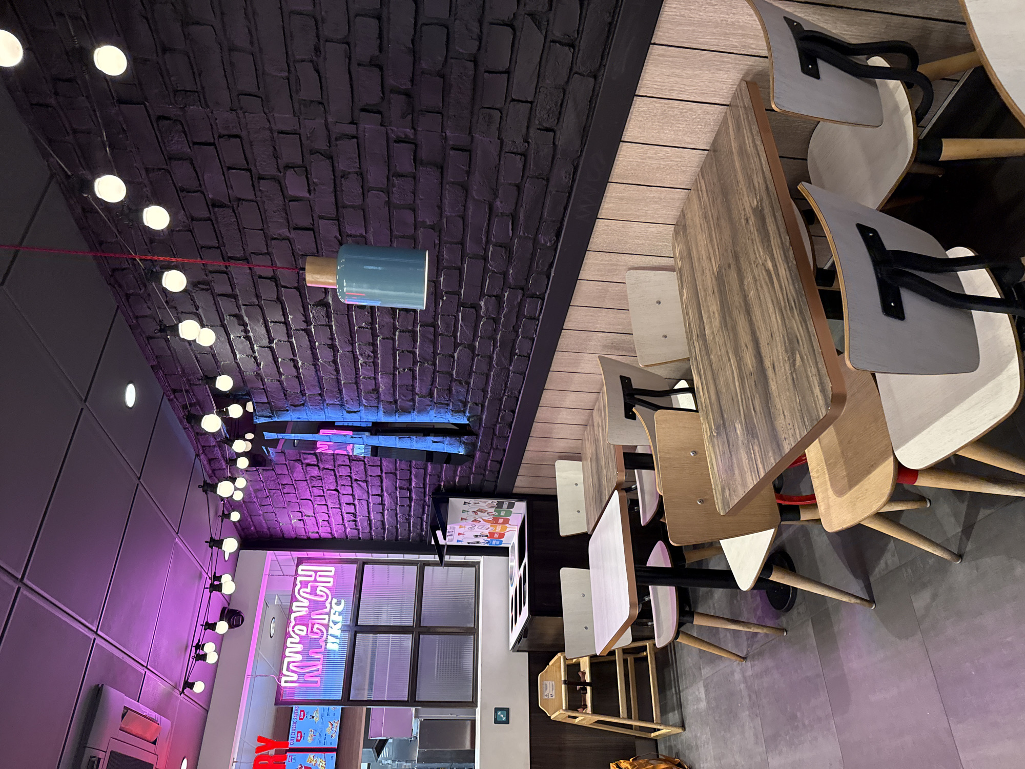 The image depicts the interior of a Krunch KFC restaurant in Gorleston-on-Sea, United Kingdom. The restaurant has a modern design with exposed brick walls, pendant lighting, and wooden tables with chairs. A neon Krunch KFC sign is visible on the wall. There is no sign of any people in the image.