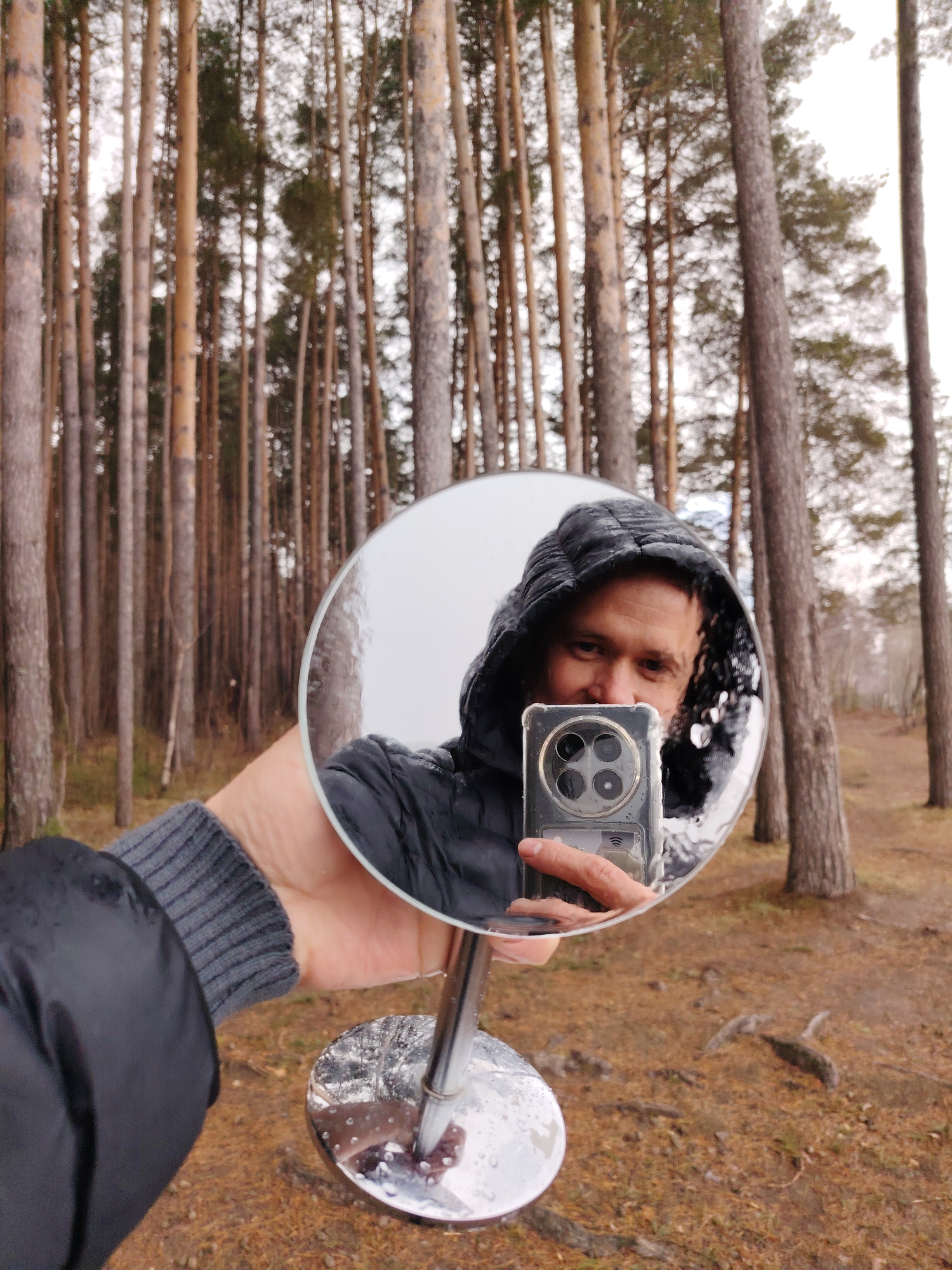 A man is taking a selfie in a forest in Chaikovsky, Russia using a round, hand-held mirror. The man is wearing a black hooded jacket, and the hood is up. He is holding a smartphone with several camera lenses in his hand, which is reflected in the mirror. The mirror's surface has water droplets on it. The man's other hand is visible, wearing a grey, ribbed-knit sleeve under the jacket. The mirror has a chrome stand with a round base. The ground is covered with brown pine needles and other forest floor debris. The background consists of tall, slender pine trees with reddish-brown bark, densely packed together, typical of a coniferous forest. The sky is overcast, creating soft, diffused lighting in the forest.