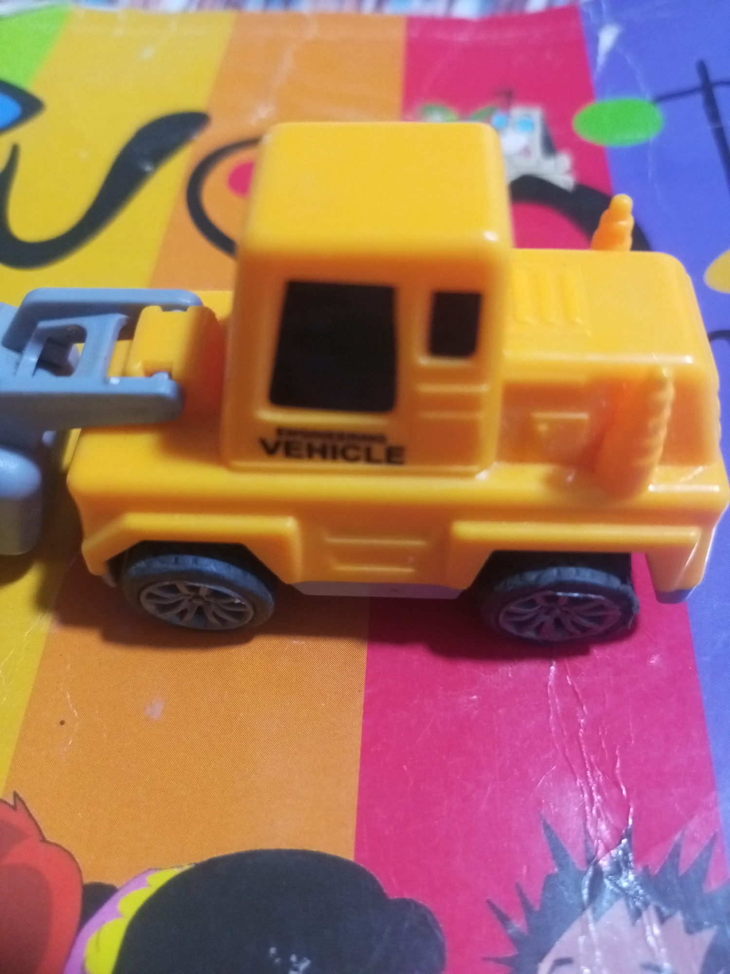 A yellow toy truck is positioned on a surface that appears to be a colorful mat or blanket. The truck has a cab with black window outlines and the text "ENGINEERING VEHICLE" printed on its side. It has two visible wheels on the right side, which are dark grey with a multi-spoke design. A grey tow hitch is attached to the front of the truck. The background is a blur of colors, including orange, pink, and purple, suggesting a patterned surface. There are no people visible in the image. The location of La Tagua, Venezuela is not discernible from the provided image.
