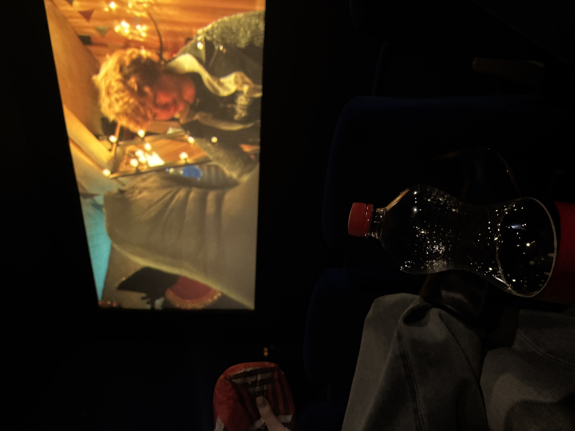 The image depicts a person sitting in a cinema, presumably in Perm, Russia, with a soda bottle and a bag of chips on their lap. A movie screen is visible on the left side of the frame, displaying a scene with a person under lights and a tent like structure. The environment is dark, typical of a movie theater setting. The focus is on the person's lap with the bottle and chips, indicating a common movie-watching experience. The cinema seats are dark blue.