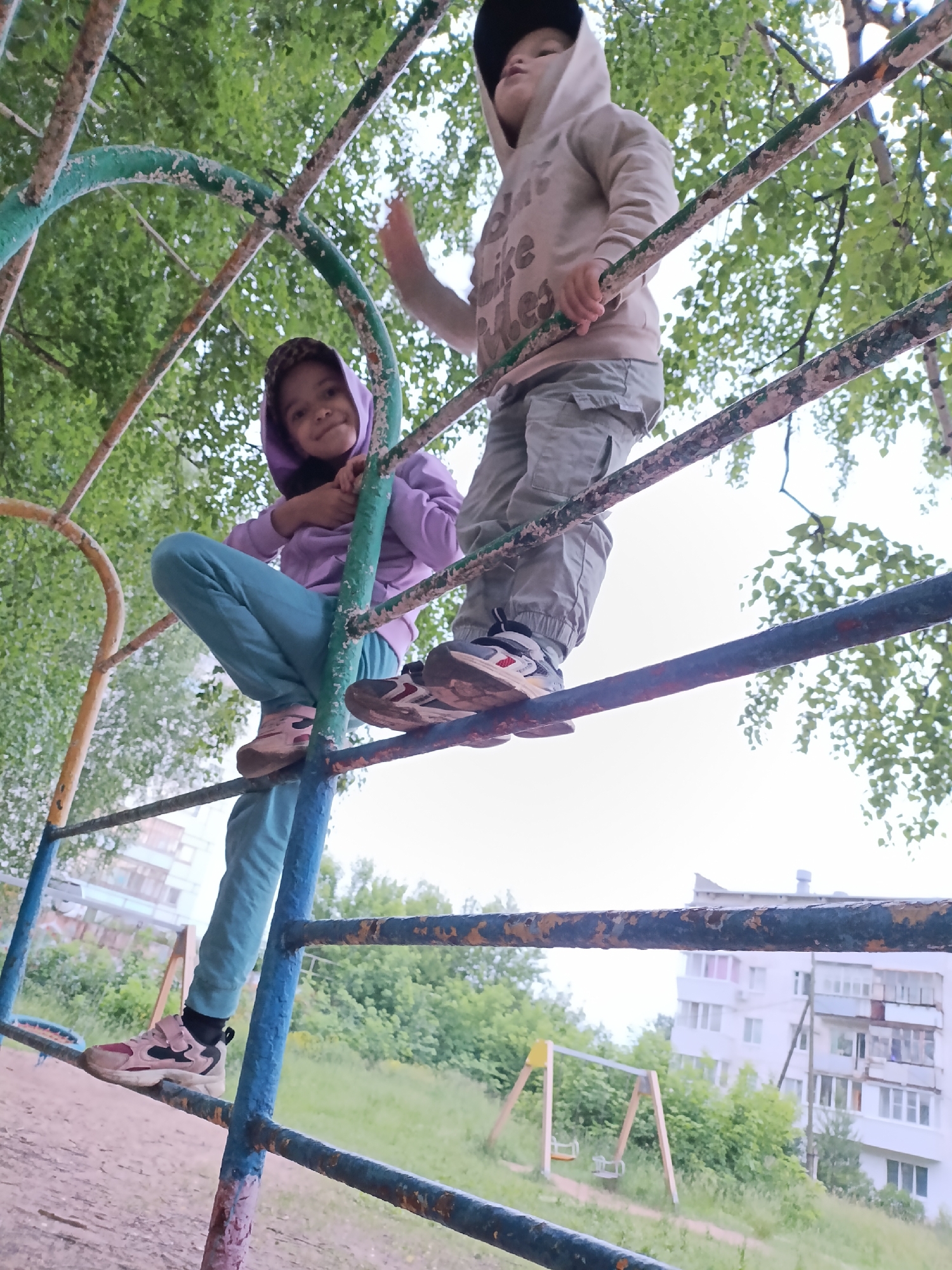 This image was taken on a playground in the city of Chaikovsky, Russia. Two children are visible on a set of monkey bars. The child on the left, a girl, is wearing a purple hooded sweatshirt and teal pants. She is sitting on one of the bars, looking at the camera with a smile. The child on the right, a boy, is wearing a beige hooded sweatshirt and gray cargo pants. He is standing on a higher bar, holding onto the one above him, and looking up and to the left. The monkey bars are made of metal and are painted in various colors, including green, blue, and yellow. Some of the paint is chipped and rusty. The background shows trees, grass, and a building. The sky is overcast.