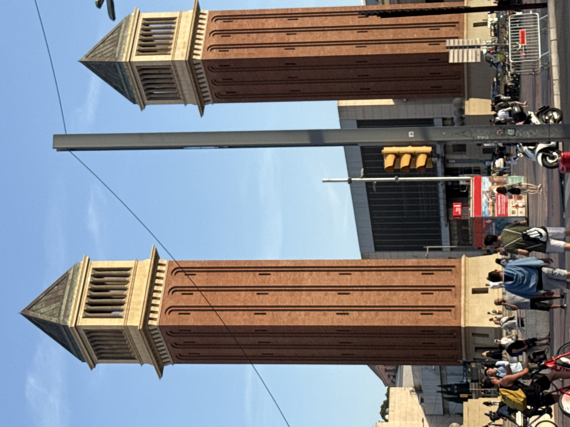 This is a daytime, eye-level, medium shot of the Venice Pavilion, two prominent red brick towers with light-colored colonnades and dark, pyramidal roofs, situated in the Plaza de España in Barcelona, Spain. The towers stand against a clear, light blue sky. In the foreground, a paved area is populated by a mix of pedestrians and cyclists. A man wearing a yellow backpack and dark clothing is on a bicycle in the lower left corner, facing right. Next to him, another bicycle is parked. A white scooter is parked in the middle ground to the right of the frame. A traffic light, showing yellow, is mounted on a gray pole in the center of the image, partially obscuring the right tower. Behind the traffic light, a modern building with dark, horizontal-pane windows is visible. Several people are walking and standing in the scene, some interacting with each other. Two towers are the central architectural elements, with a metal pole and wire running diagonally through the frame from the upper left to the lower right.