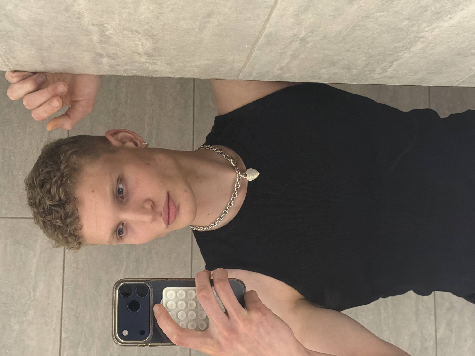 A young, light-skinned person with short, curly light brown hair and blue eyes is captured in a mirror selfie. They are wearing a black ribbed sleeveless top and a silver-toned chain necklace with a small, heart-shaped pendant. A silver hoop earring is visible in their left ear.
The person holds a dark-colored smartphone, consistent with an iPhone, in their right hand. The phone is encased in a clear or dark-framed case that features a white, grid-patterned accessory on its back, which a finger is resting upon. The person's left hand is visible in the upper left of the frame, seemingly resting on a ledge or the top edge of a tiled surface.
The background and immediate setting consist of light grey, possibly stone-look, tiled surfaces, suggesting an indoor environment such as a bathroom or modern room. The person's expression is neutral. The text on the heart-shaped pendant is not clearly legible. The photo is taken from a slightly elevated perspective, looking down into the mirror.