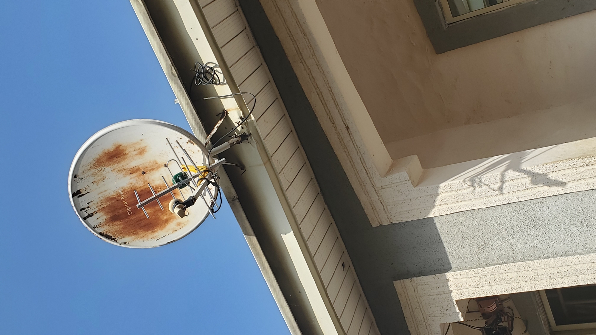 A rusted satellite dish and a TV antenna are mounted on the eave of a building under a clear blue sky. The satellite dish is white with large areas of rust and some black streaks. The antenna is silver with several horizontal bars. Wires are attached to the antenna and extend downwards. The building has a light-colored soffit with horizontal slats and a gray textured wall. Decorative white trim frames a window on the lower left of the image. The image was taken from a low angle looking up at the dish and antenna.