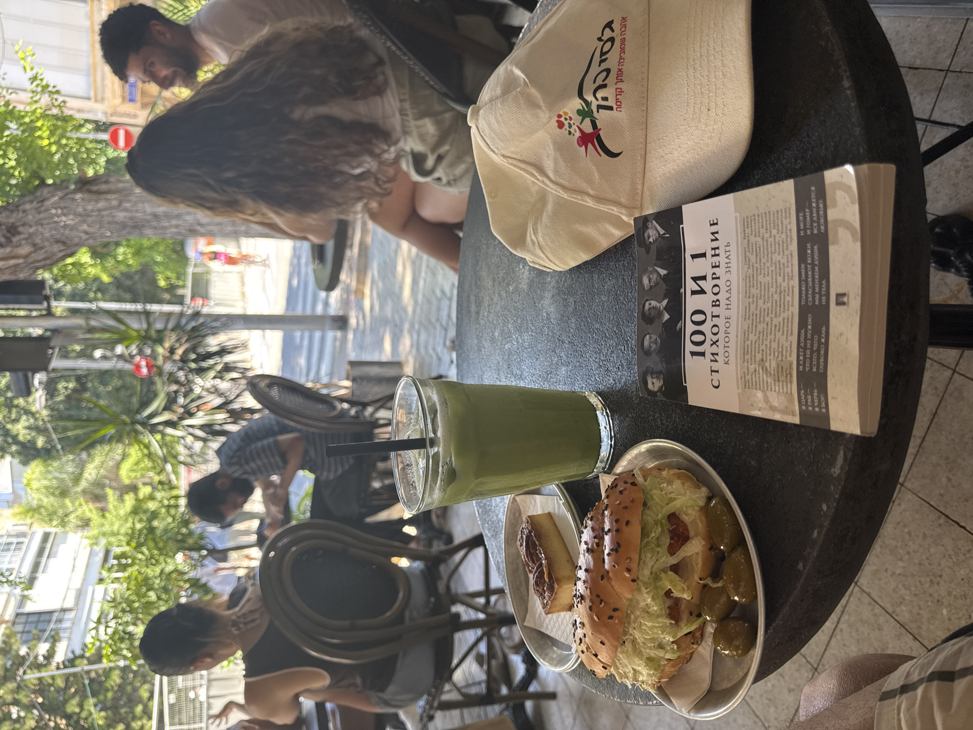 The image depicts a table at an outdoor cafe in Tel Aviv, Israel. In the foreground, a sandwich, a small rectangular pastry with pecans, and a glass of green juice with a straw are arranged on a dark, textured surface. To the right of the food items, a beige baseball cap with a colorful logo and Hebrew text is placed. A book with a black and white cover featuring portraits of several men and Hebrew text is also on the table. In the background, blurred figures of people are seated at tables and walking, suggesting a busy cafe environment. Trees and buildings are visible, hinting at an urban setting. The lighting suggests it is daytime, possibly a sunny afternoon.