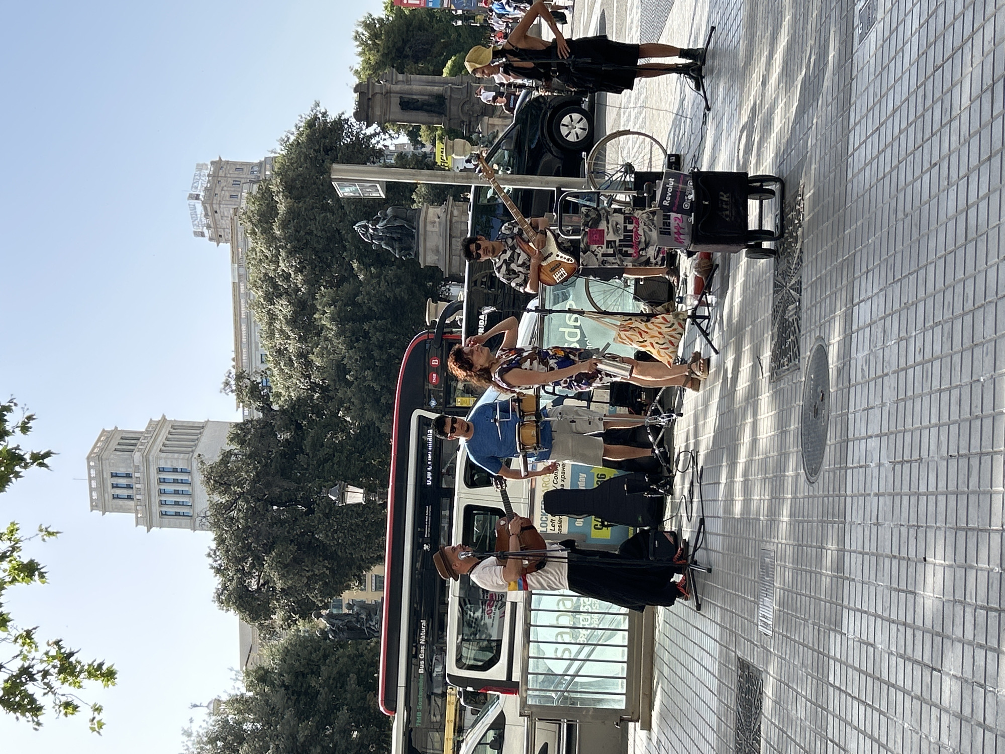 In Barcelona, Spain, on a sunny day, a group of street musicians performs in front of a crowd. The musicians include a man playing a guitar and singing, another man playing drums, a third man playing a bass guitar, and a woman singing. They are set up on a tiled pedestrian walkway, with a bus and several cars visible in the background. In the distance, large buildings and trees can be seen. The atmosphere is lively and vibrant, with people enjoying the music and the warm weather. The scene captures a typical day in the city, with its bustling streets and cultural offerings.