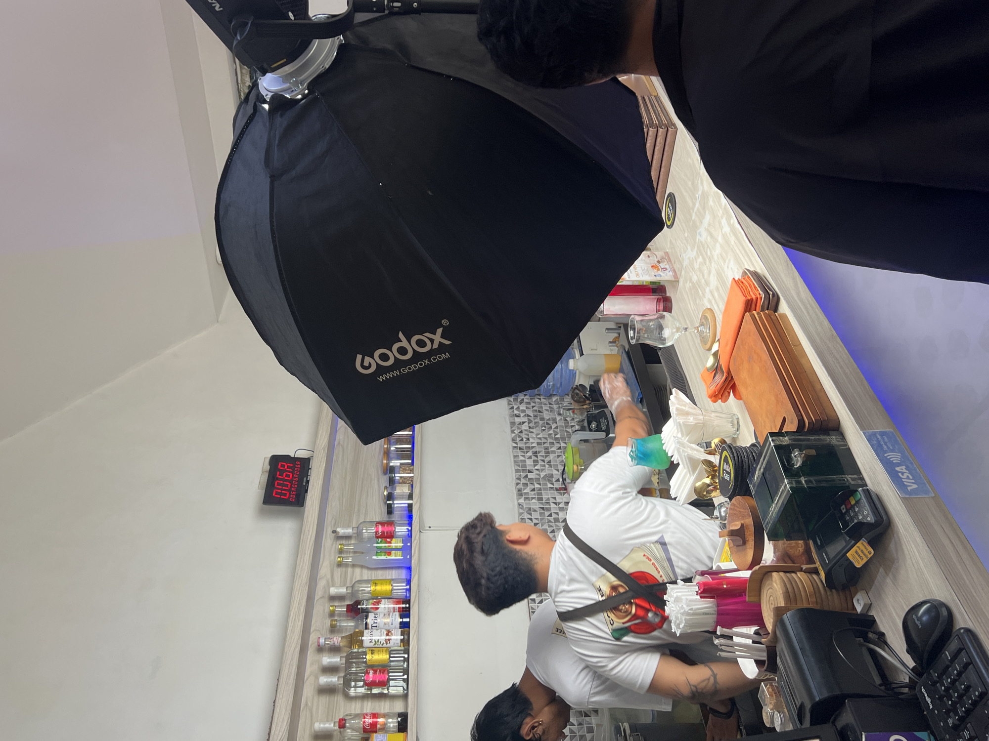The image shows the interior of what appears to be a bar or restaurant. A man is working behind a counter, likely preparing drinks. A Godox brand softbox lighting setup is positioned overhead. Various bottles are arranged on shelves behind the counter. A digital display shows "0068" indicating possibly the time. A Visa sign is visible on the counter. The location is likely in Sukawati, Indonesia, as per the prompt's instructions.