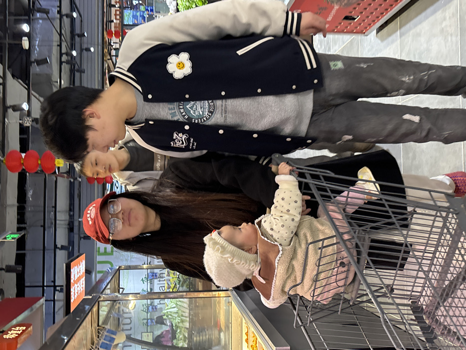 A group of four individuals, three adults and one infant, are observed inside a supermarket in Jiangjin, China. In the foreground, an infant is seated in the child's seat of a metal shopping cart, facing right. The infant wears a cream-colored knitted hat with ruffles, a light brown and cream-patterned jacket, and pink pants. Behind the cart and to the left stands a woman with long dark hair, wearing a red baseball cap, glasses, a dark long-sleeved top, and light-colored pants with red footwear. She looks slightly upward and to the right. To her right, a man in a navy blue and cream varsity-style jacket with a white daisy patch featuring a yellow smiley face, a grey t-shirt, and dark, distressed pants with paint splatters, is looking towards the woman. Behind him, another man with short dark hair is partially visible, wearing a light-colored jacket over a dark t-shirt, looking forward. The supermarket environment features display cases, including one with a glass front containing what appears to be baked goods or prepared food on the left. Fresh produce is visible on shelves in the background. Red lanterns hang from the ceiling, which is fitted with fluorescent lighting. Green and white signage, including the partial text "ENJE" and "healthy" in English alongside Chinese characters, is visible. The floor is tiled.
