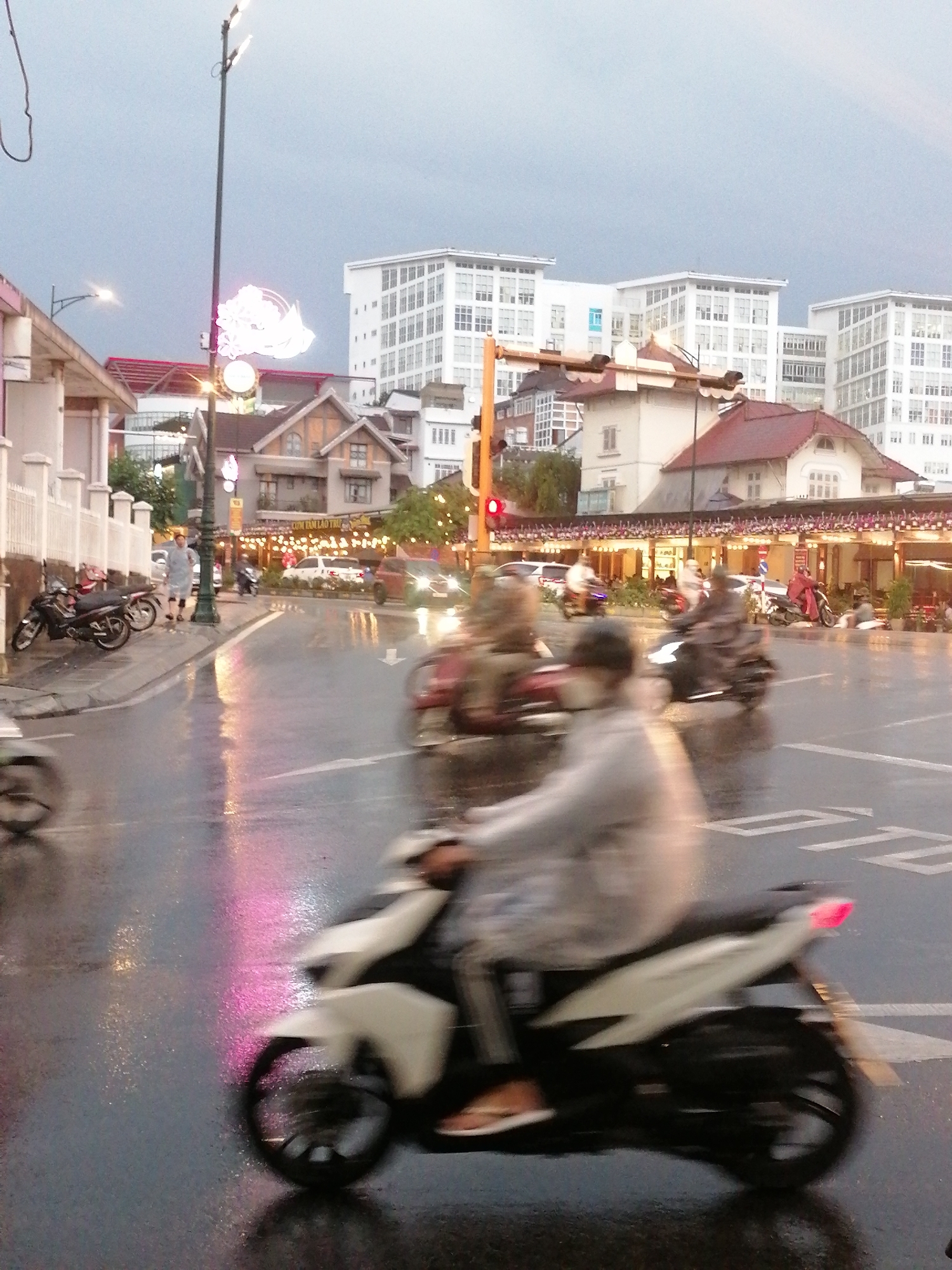 The image shows a wet street scene in Da Lat, Vietnam, likely after a rain. Motion blur indicates active traffic. A person on a white scooter drives in the foreground. Several other motorbikes are present in the street. Buildings line the street. There is a traffic light. Street lights are present, and there appears to be a decorative light fixture resembling a floral or avian form mounted on a pole. People are walking on the sidewalk. There are parked motorbikes along the curb. The general light suggests it is either dusk or a heavily overcast day.
