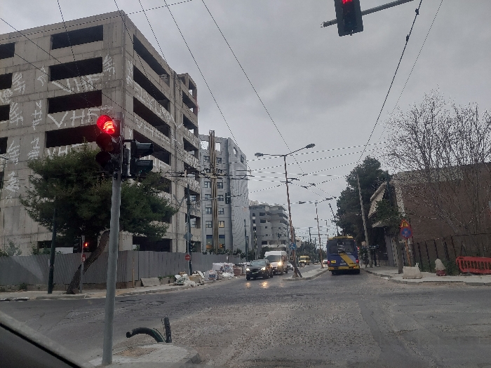 This image depicts a street scene in Kallithea, Greece, under an overcast sky. The perspective is from inside a vehicle at an intersection, looking down a multi-lane asphalt road that appears damp.
In the immediate foreground on the left, a grey traffic light pole is prominent, displaying an illuminated red signal. Behind this, a small green tree partially obscures the lower section of a large, multi-story concrete building, which appears unfinished or abandoned. This building features multiple rectangular open windows/balcony spaces and is extensively covered in white graffiti. Along the base of this building, a temporary metal fence separates it from the sidewalk, where piles of construction debris and white bags are present.
Overhead, a dense network of black utility wires and cables crisscrosses the sky, supported by numerous streetlights and poles extending down the road.
Further down the street, several vehicles are observed moving away from the viewer. Two cars in the center lane have their headlights illuminated. To the right, a yellow and blue public transport vehicle, likely a bus or trolleybus, is also proceeding down the road.
On the right side of the street, a dark wall or fence runs parallel to the road, with bare-branched trees visible behind it. A yellow triangular warning sign and a purple circular sign are affixed to a pole on the sidewalk. Red and white plastic traffic barriers are also present on the right-hand sidewalk. In the background, additional multi-story buildings of varying architectural styles are visible. No explicit individuals are discernible in the scene.