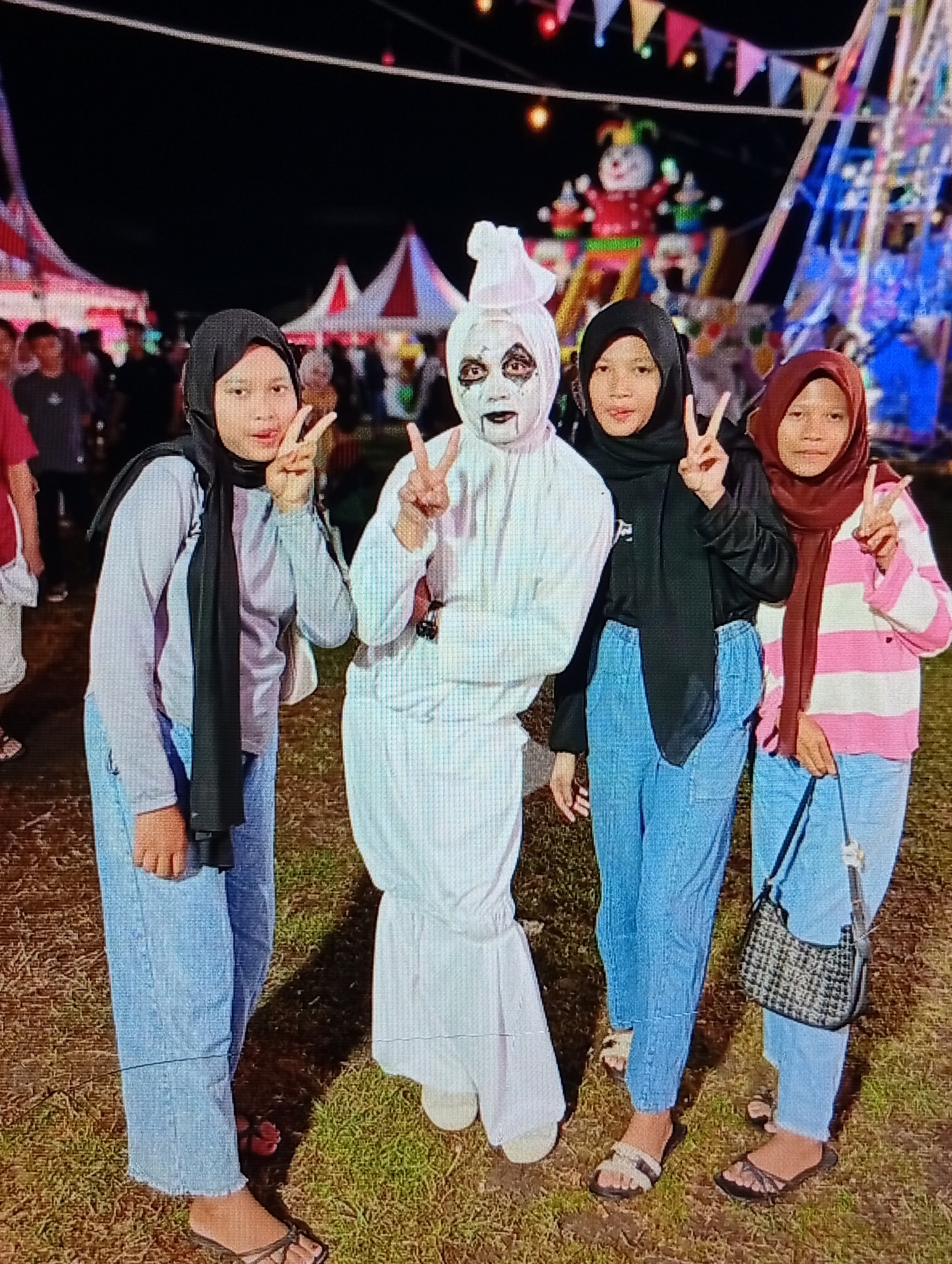 Four individuals, appearing to be young females, are posing for a photograph at a nighttime outdoor event, identified as being in Lhokseumawe, Indonesia. The setting features carnival-like elements including red and white striped tents, an illuminated inflatable clown-like figure, colorful bunting overhead, and a large, brightly lit amusement ride, possibly a Ferris wheel, in the background. The ground is dry grass with visible soil.
From left to right:
1. A female wearing a light grey long-sleeved shirt, light blue jeans, and a black hijab. She is wearing dark sandals and holds her right hand up in a peace sign.
2. An individual in a full white costume, including a hooded headpiece, with their face painted white and dark around the eyes and mouth, resembling a horror character. A conical white accessory is on top of the headpiece. This person also holds their right hand up in a peace sign.
3. A female wearing a black long-sleeved shirt, light blue jeans, and a black hijab. She is wearing dark sandals and holds her right hand up in a peace sign.
4. A female wearing a pink and white horizontally striped long-sleeved shirt, light blue jeans, and a reddish-brown hijab. She carries a small black and white checkered shoulder bag on her left side and is wearing dark sandals. She holds her right hand up in a peace sign.
All four subjects are facing the camera and exhibit similar poses, indicating a planned group photo. The scene is lit by artificial lights from the carnival attractions, casting shadows on the ground.