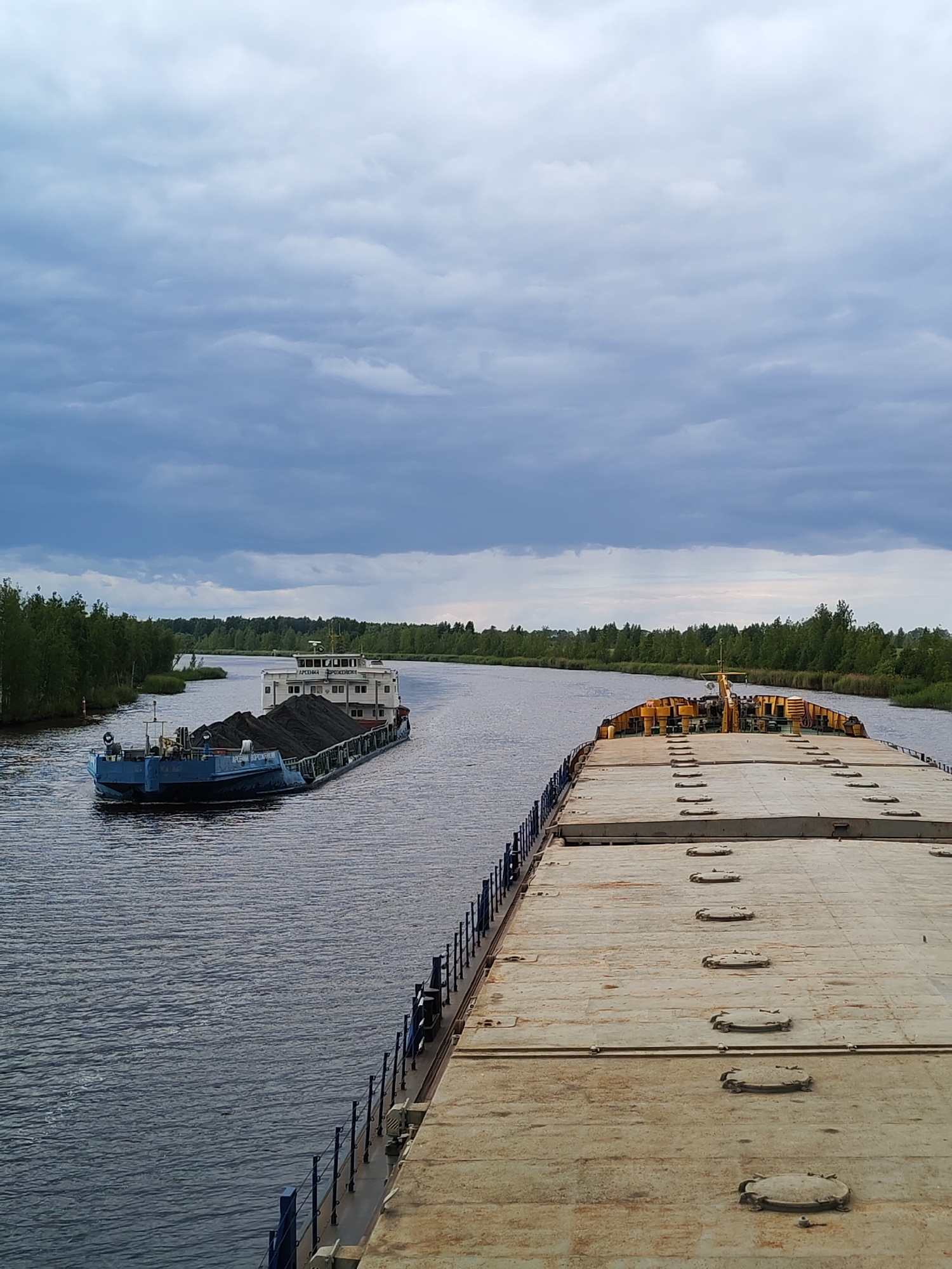 An overcast day in Ozernoe Ust'e, Russia, features a wide, calm river with two barges. In the distance, a blue barge, loaded with dark material resembling coal, navigates the waterway. Its superstructure is white, and it has a small red buoy to its left. In the foreground, the deck of a larger, lighter-colored barge stretches across the frame, revealing numerous circular hatches and yellow safety railings. The banks of the river are lined with dense green trees, under a sky filled with heavy, gray clouds.