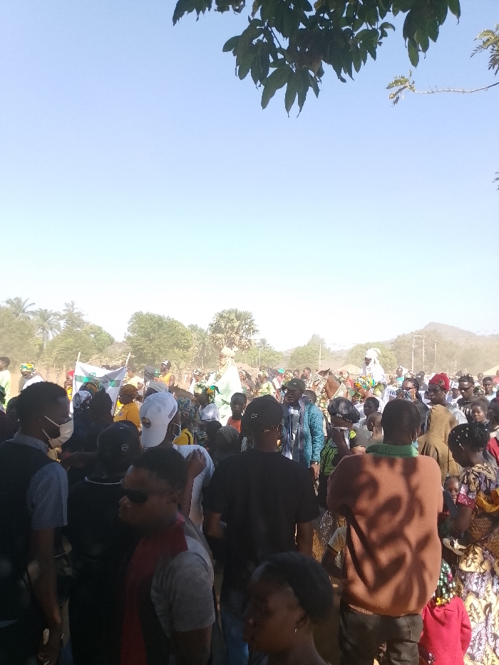 This image depicts a dense public gathering in Kpak, Nigeria, under a clear, light blue sky during daylight hours. A large crowd of individuals of various ages is tightly packed, facing forward or towards a central point. Overhead, leafy branches are visible in the top right and left corners, with additional trees, including palm trees, in the mid-ground. Distant hills are visible on the horizon to the right. The ground appears dusty, contributing to a slight haze in the background.
Two individuals are prominently featured on horseback, elevated above the surrounding crowd. The person on the left rides a light-colored horse and is dressed in light-colored, possibly white or light green, traditional attire with a matching head covering. The person on the right rides a darker horse and also wears light-colored attire with a head covering.
Among the crowd, several individuals are wearing face masks. Attire is diverse, including traditional patterned clothing and casual wear. A white flag with a green emblem or text is visible, held upright to the left of the horse riders. A green flag is partially visible further right in the crowd. The scene indicates a public procession or outdoor event.