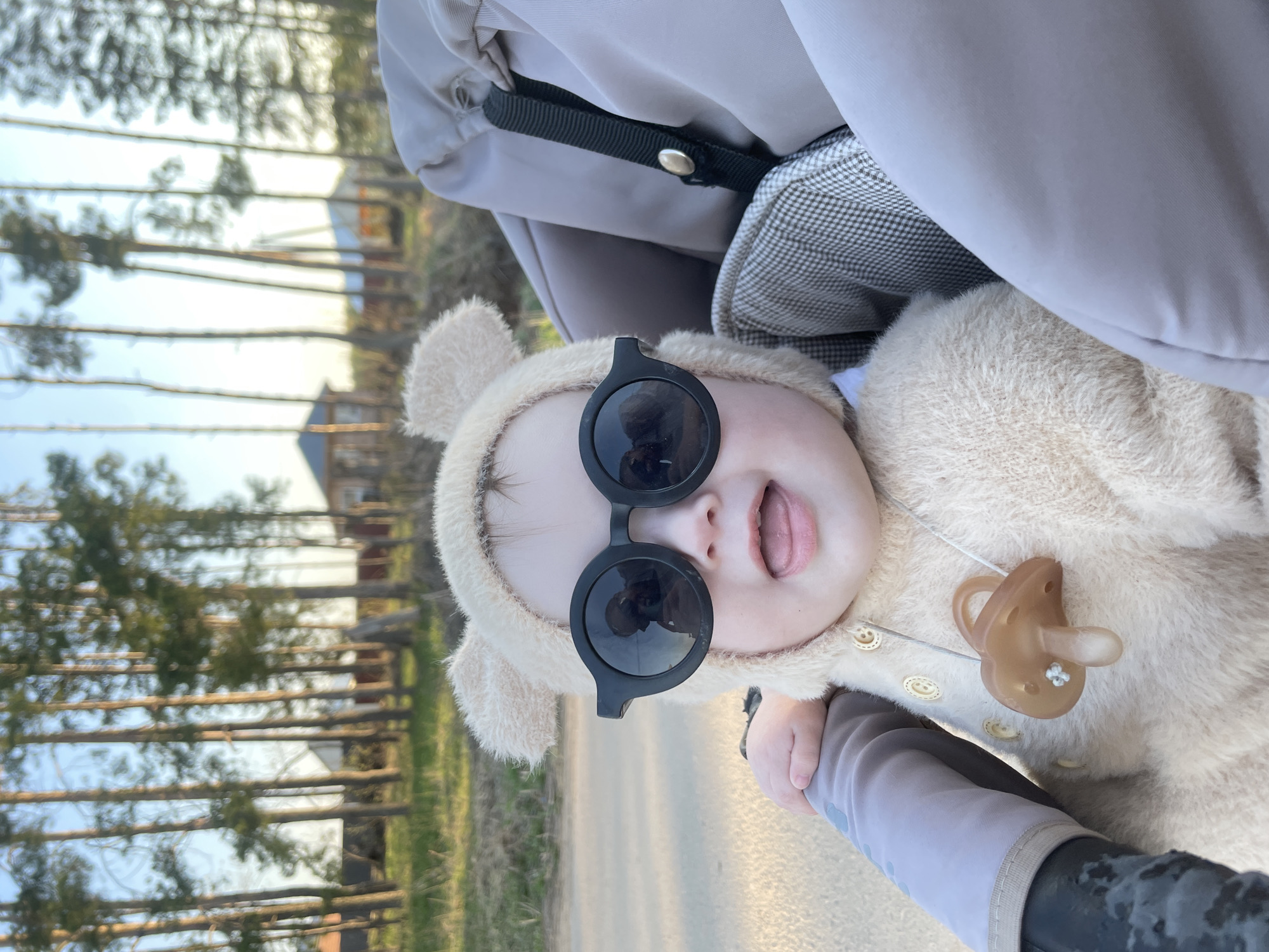 A baby wearing sunglasses and a furry bear costume sits in a stroller. The baby's mouth is open, revealing their tongue. The stroller has a grey and black patterned interior and a beige exterior. In the background, there are trees and a building. The location is Подшивалово, Russia.