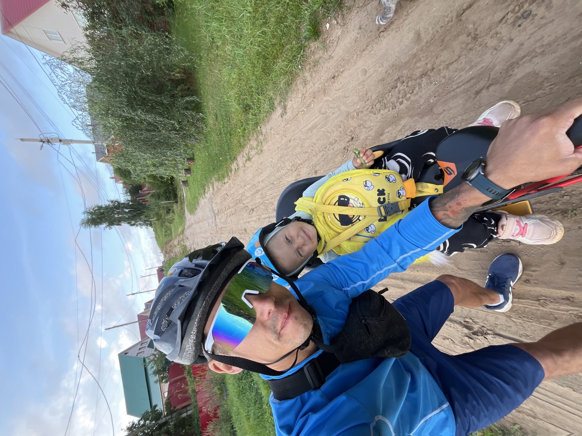 The image depicts a man and a young child riding a bicycle in Chaikovsky, Russia. The man is in the foreground, wearing a blue cycling jersey, blue shorts, a black helmet, and mirrored cycling glasses. He is holding the handlebars of the bicycle. The child is seated in a child seat on the back of the bicycle, wearing a yellow jacket and a blue helmet. The child is smiling and holding a green stalk. The background shows a dirt road with grass and trees on either side, and houses with red roofs in the distance. The sky is cloudy. A cat is partially visible on the right side of the frame. The setting appears to be a rural or suburban area.