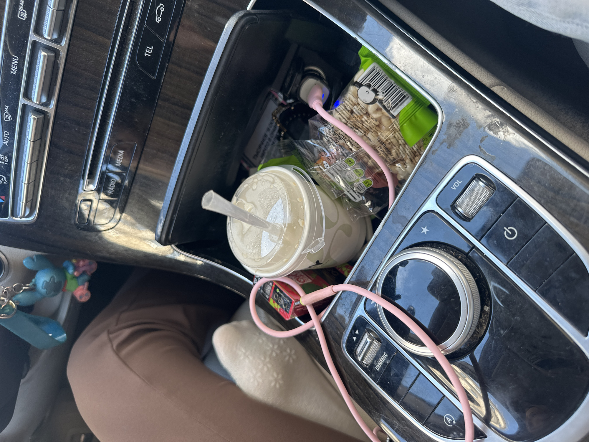 The image shows a close-up view of a car's center console, cluttered with various items. The console features buttons and a rotary dial, suggesting it is from a modern vehicle. Among the objects are a disposable cup with a straw, a bag of snacks, a charging cable, and what appears to be a packet of gum or candy. A pink charging cable is plugged into a car charger, powering a device. The overall scene suggests a person is in their car, possibly on a trip or during their daily commute, with snacks and a drink readily available. The presence of a pink charging cable and a small blue stuffed toy keychain hanging from the dashboard hints at a personal touch and potentially a female owner. The car's interior appears somewhat worn, with dust visible on the console. The lighting suggests it is daytime, but the specific time is not determinable. The text visible on the packaging is in Cyrillic script, with some words appearing to be in Russian and Kazakh. The text includes brand names and nutritional information. No people are visible in the frame.