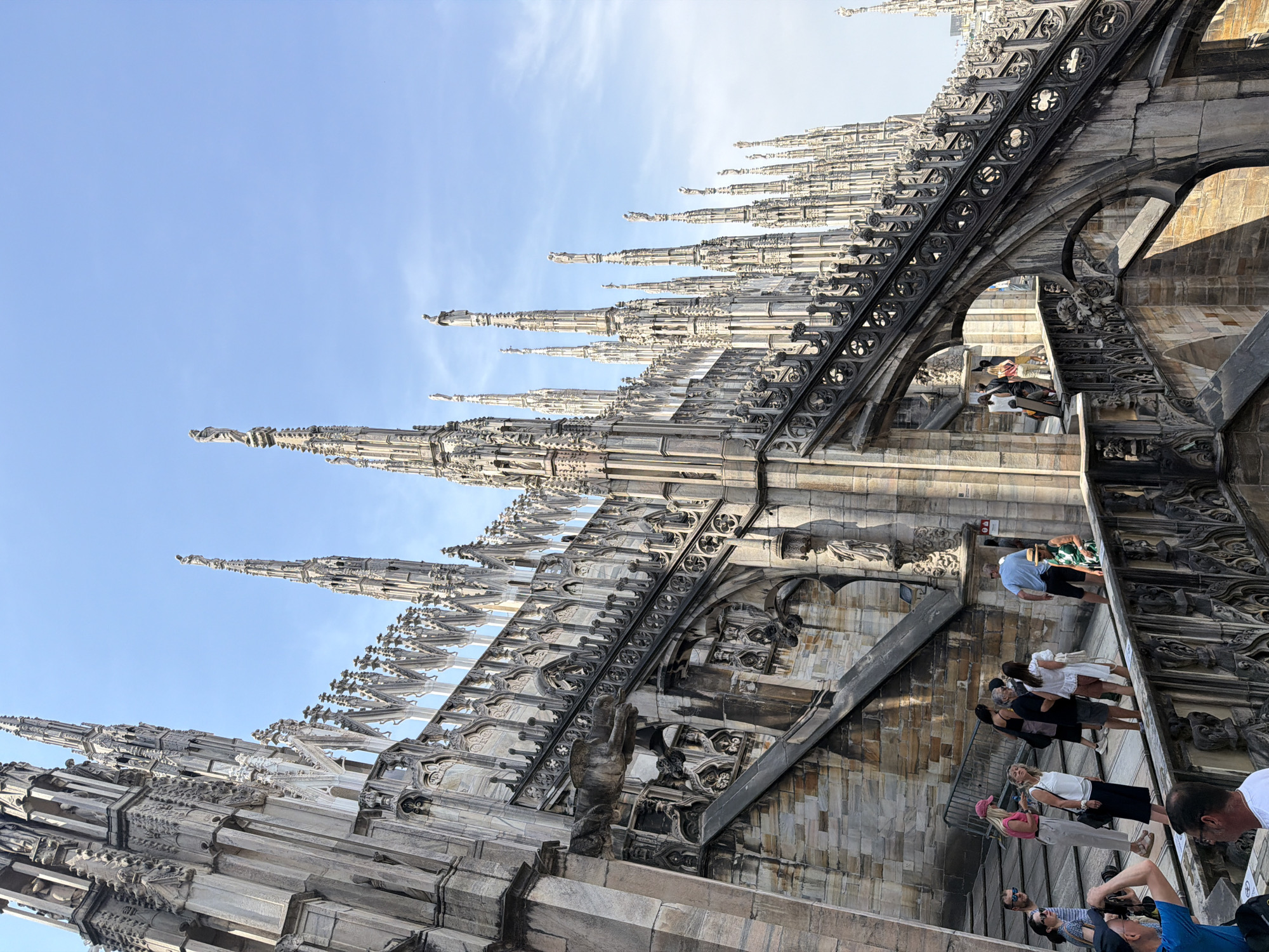 The image captures the rooftop of the Milan Cathedral (Duomo di Milano) in Milan, Italy. The architecture features numerous spires and Gothic ornamentation. A group of tourists is visible walking along the rooftop, taking pictures and observing the surroundings. The sky is blue with some wispy clouds. The rooftop surface is constructed from light-colored stone blocks.