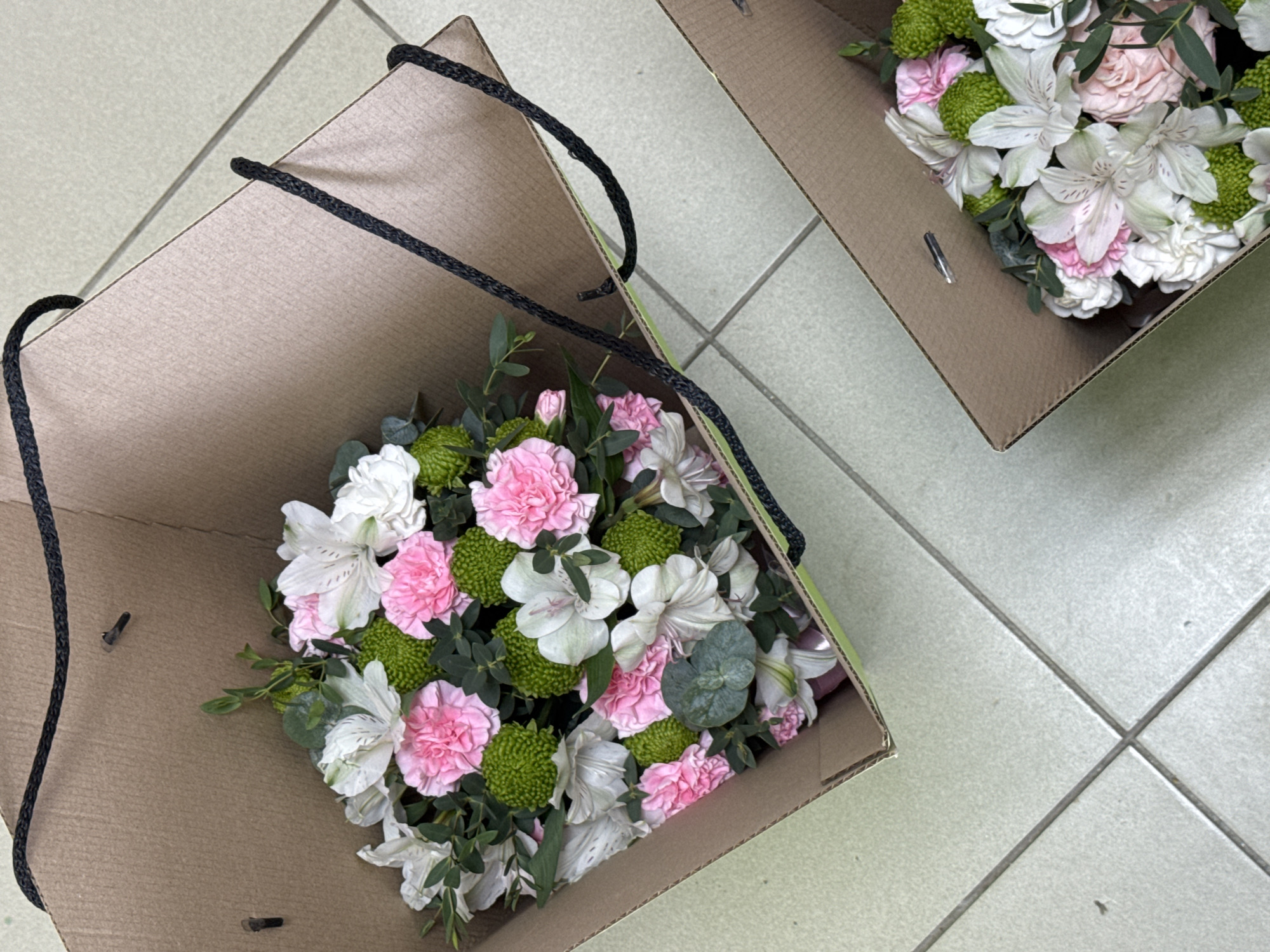 The image shows two cardboard boxes filled with bouquets of flowers. The box on the left is closer to the viewer and has black rope handles. The flowers in the box include pink carnations, white alstroemeria, and green chrysanthemums, along with green foliage. The box on the right is further away and also contains flowers, but the specific types are less distinct. The scene is indoors, likely on a tiled floor. No people are visible in the image. There is no specific information about the location in Izhevsk, Russia, in the image.