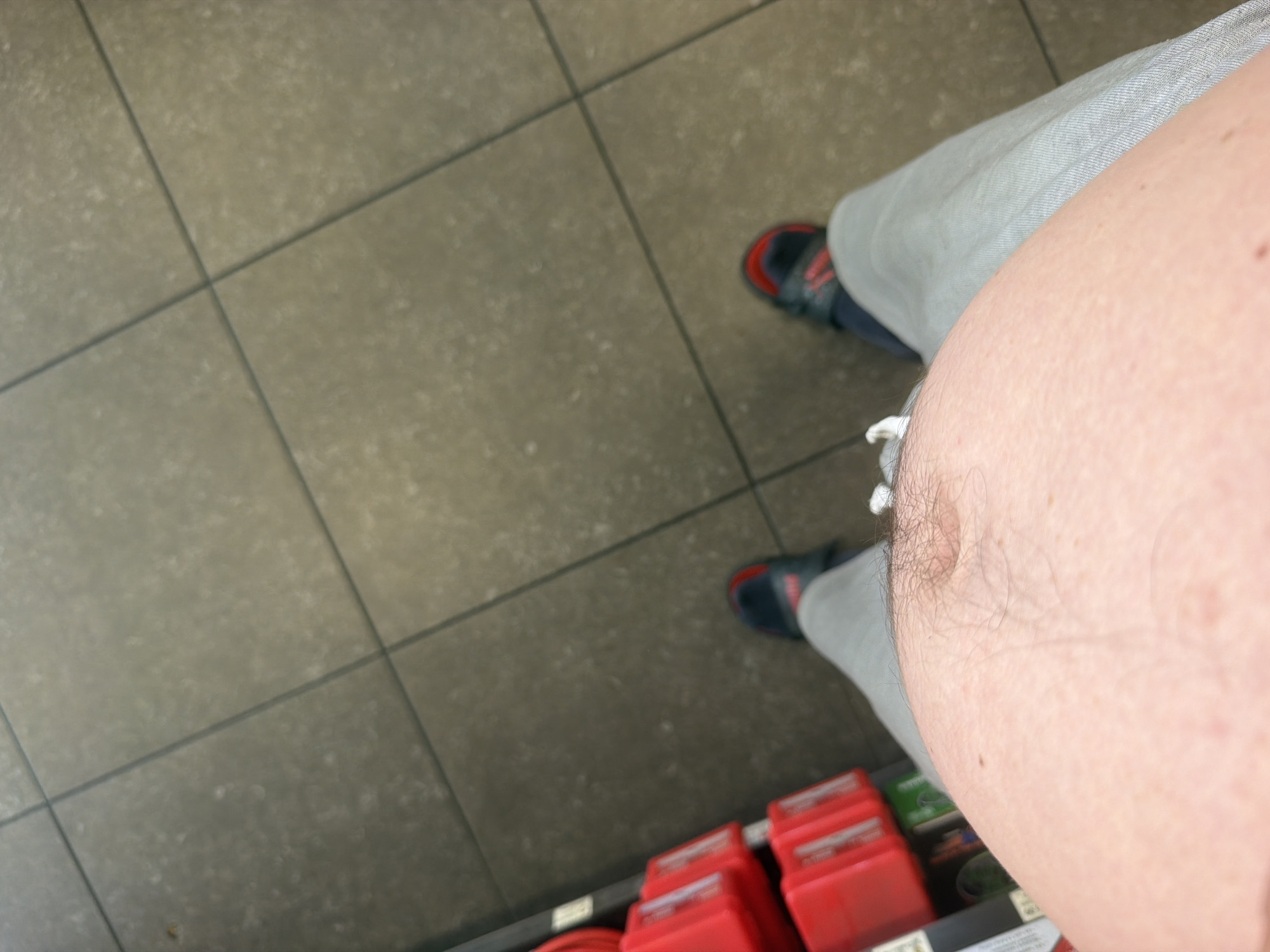 The image is a close-up, first-person perspective shot of a person's torso and feet, standing on a tiled floor. The person is wearing light gray sweatpants and dark athletic shoes with red accents. To the left of the frame, there are several red storage containers stacked on shelves, suggesting an indoor environment like a store or warehouse. The floor tiles are large and a muted brown or gray color, laid in a grid pattern. There is no visible indication of the city or country.