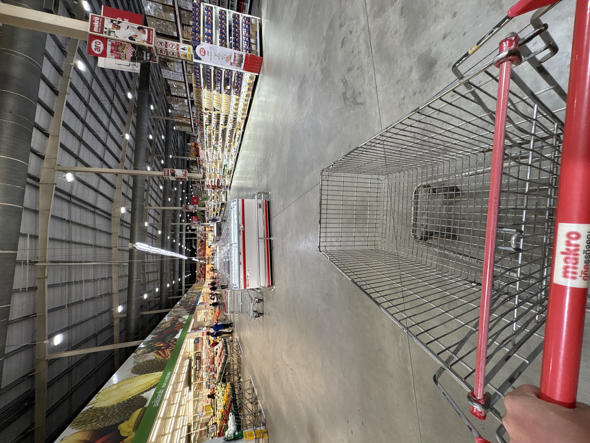 The image, taken inside what appears to be a Makro store in Baan Bo Phut, Thailand, presents a first-person perspective from someone holding a shopping cart. The cart is made of metal wire with a red handle bearing the Makro logo and Thai text. The scene stretches down a long aisle lined with shelving units stocked with various goods.
On the left, the shelving units begin with fresh produce like durian, transitioning to other items. The right side features refrigeration units and aisles filled with packaged goods. Signage above the shelves displays the "aro" and "Makro" brands, along with details about Italian food.
The store's architecture includes a high ceiling with exposed beams and numerous overhead lights. People can be seen further down the aisle, near the refrigerators. The floor is a smooth, gray concrete.