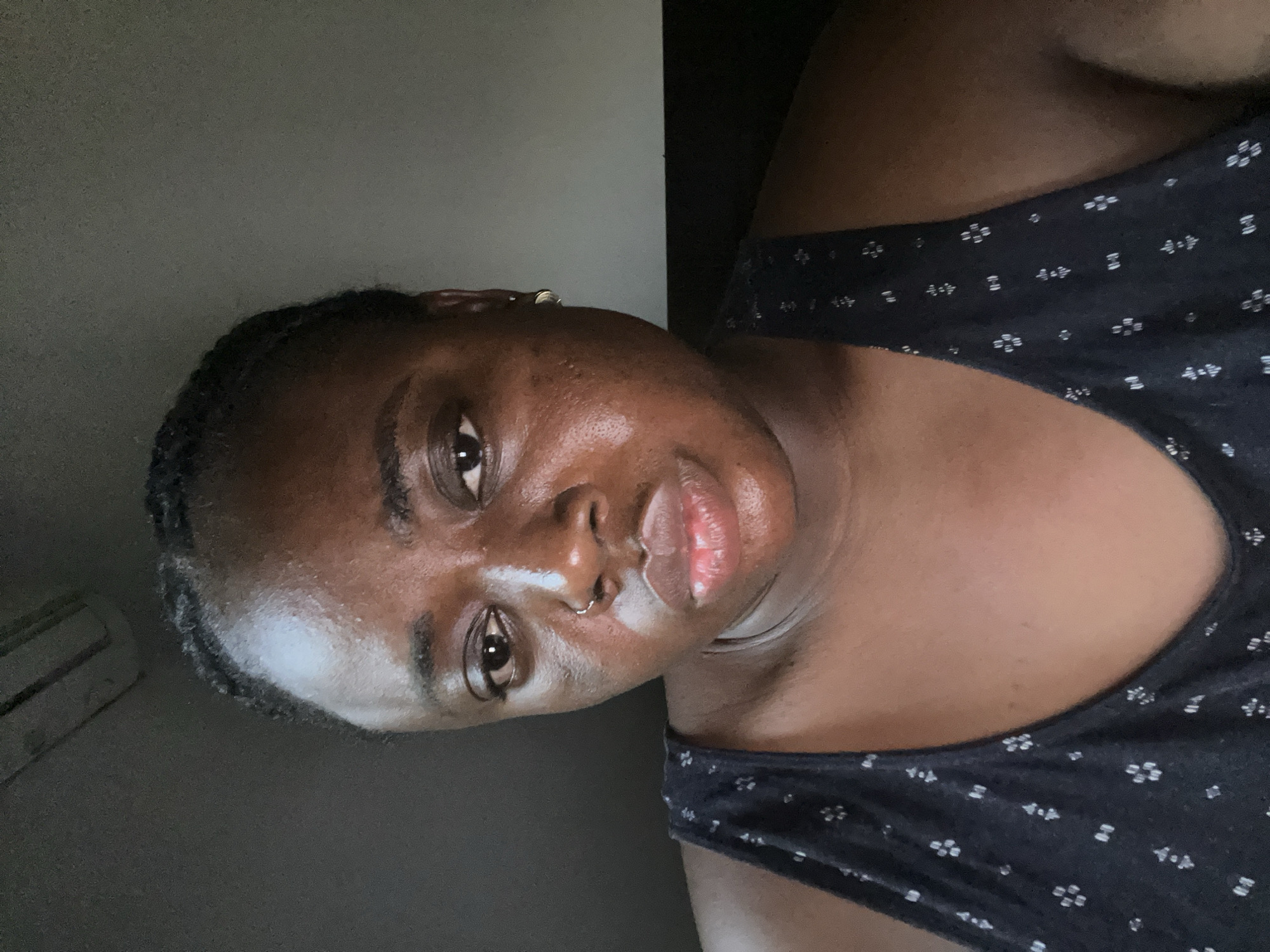 A dark-skinned woman is seen from the chest up. She is wearing a black top with small white floral patterns. Her hair is braided. She has a nose ring and an earring. The background is a neutral gray. The location is not discernible, so Abuja, Nigeria is unknown.
