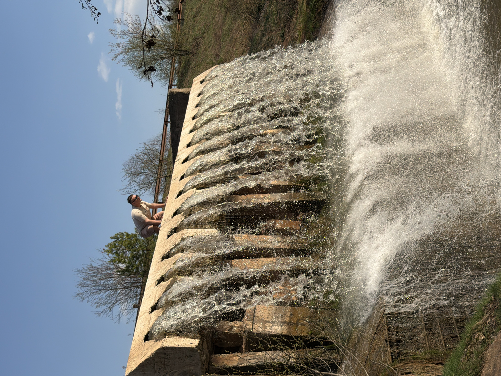 A man sits on the side of a tiered dam or waterfall in Kamennoye, Russia. The water cascades down the concrete structure, creating a misty effect. The scene is bathed in sunlight, and a blue sky is visible in the background. Vegetation flanks the edges of the dam.